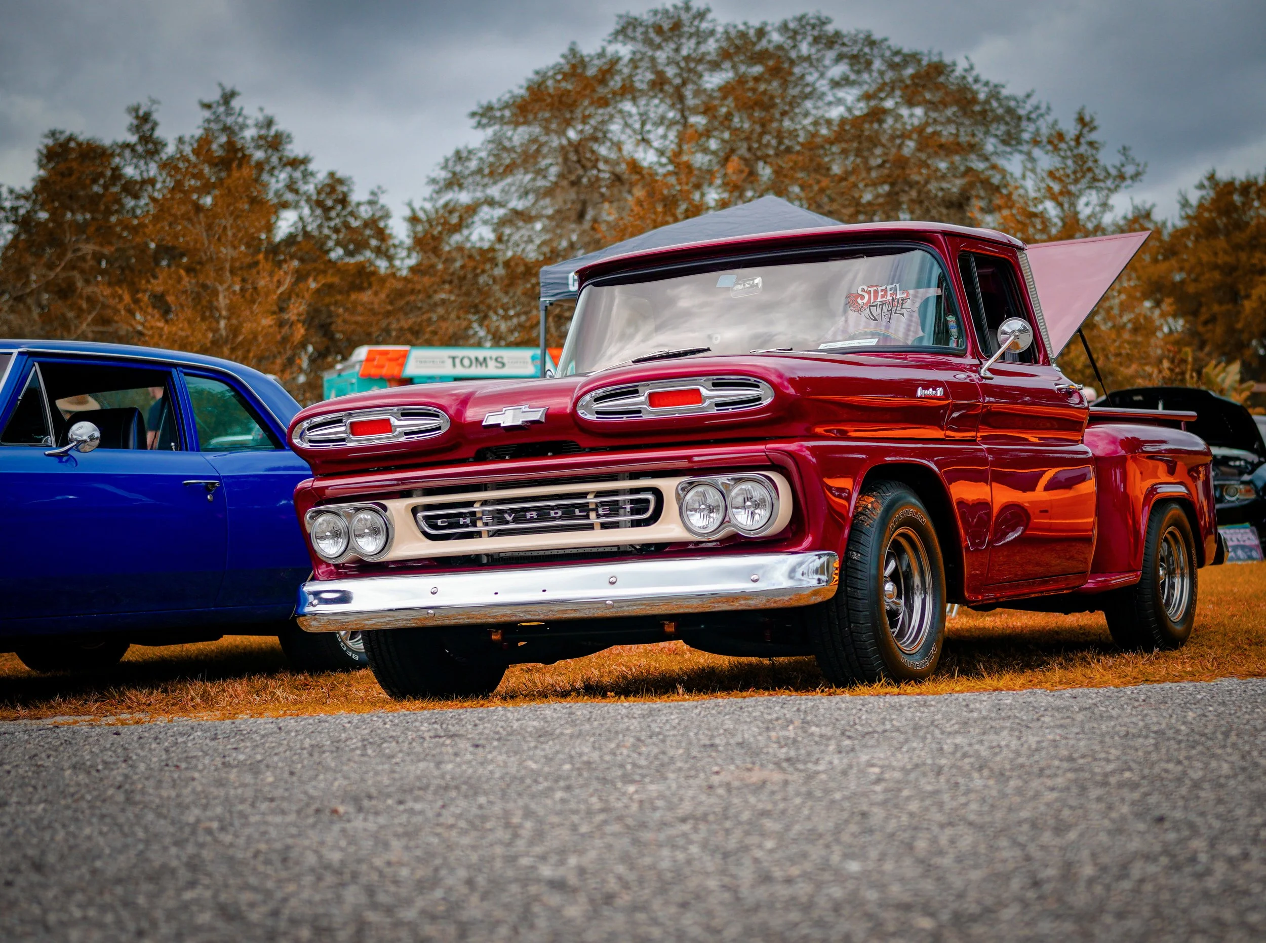 A vintage red Chevrolet truck at an outdoor car show, parked beside a blue car, with trees and a cloudy sky in the background.