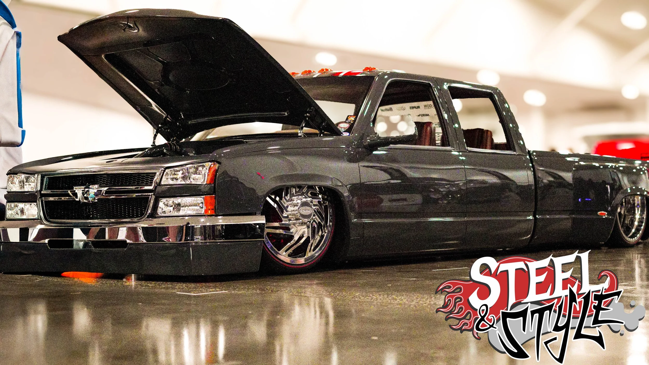 A black modified lowrider car with open hood displayed at an indoor car show, with a "Steel & Style" logo in the bottom right corner.