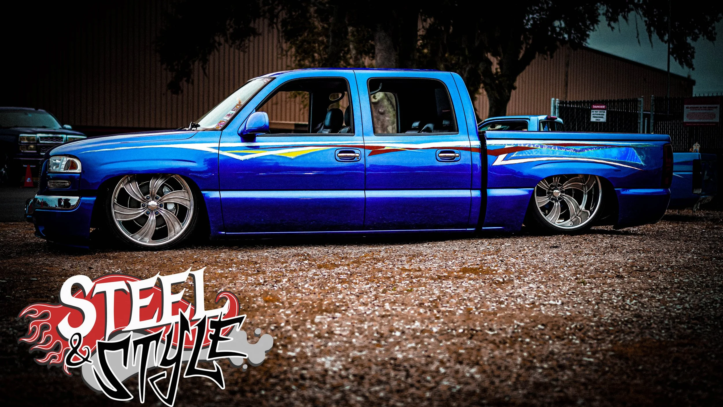 A lowered, customized blue pickup truck with large chrome wheels and flame decals parked on gravel at night. The background shows trees and fences.