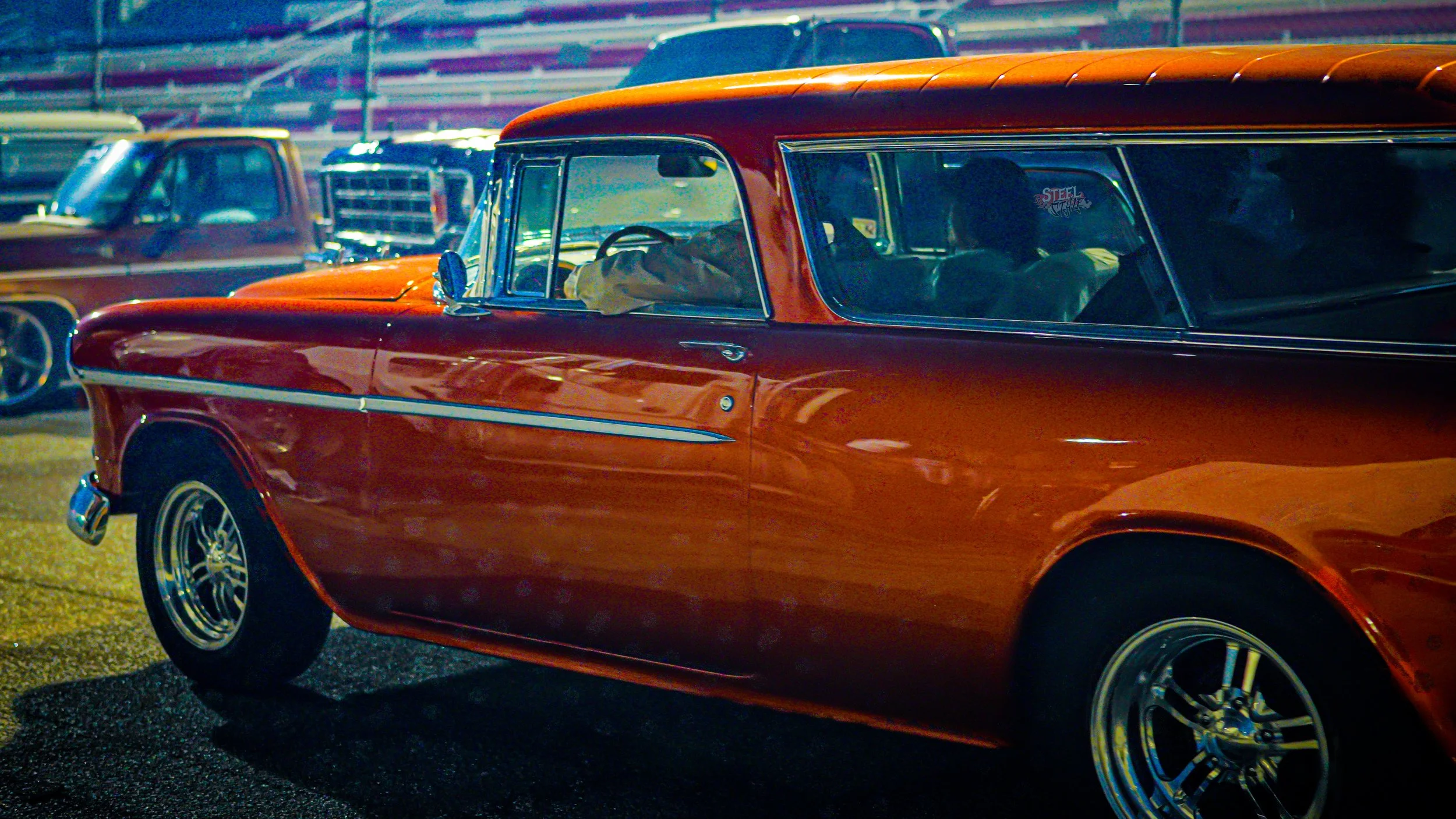 A vintage orange station wagon parked at night with other classic vehicles in the background, illuminated by artificial lighting.