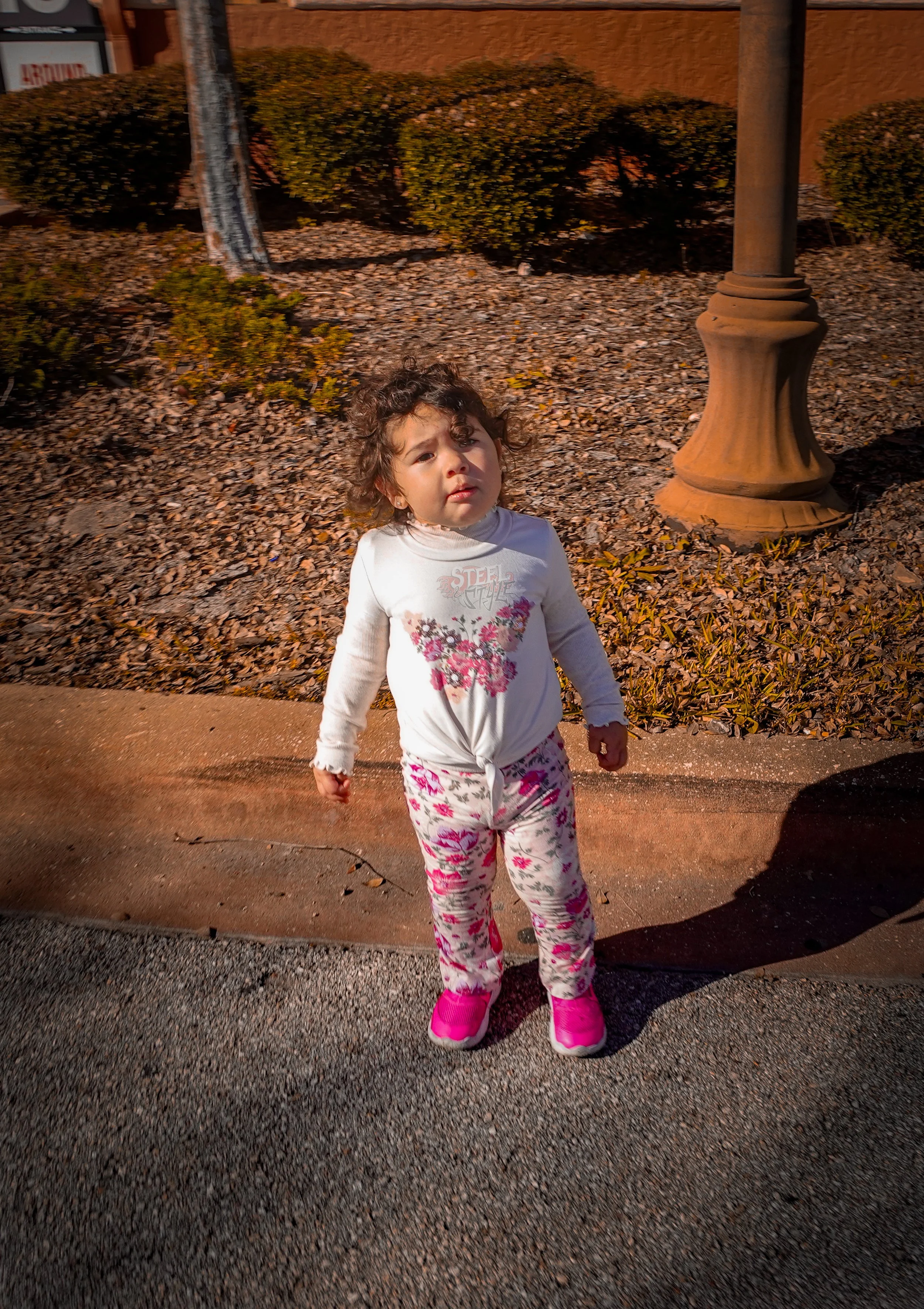 A young girl with curly hair, wearing pink sneakers, patterned pants, and a long-sleeve shirt, stands outdoors on a sidewalk in a park-like setting with bushes, gravel, and a lamppost during late afternoon or early evening.