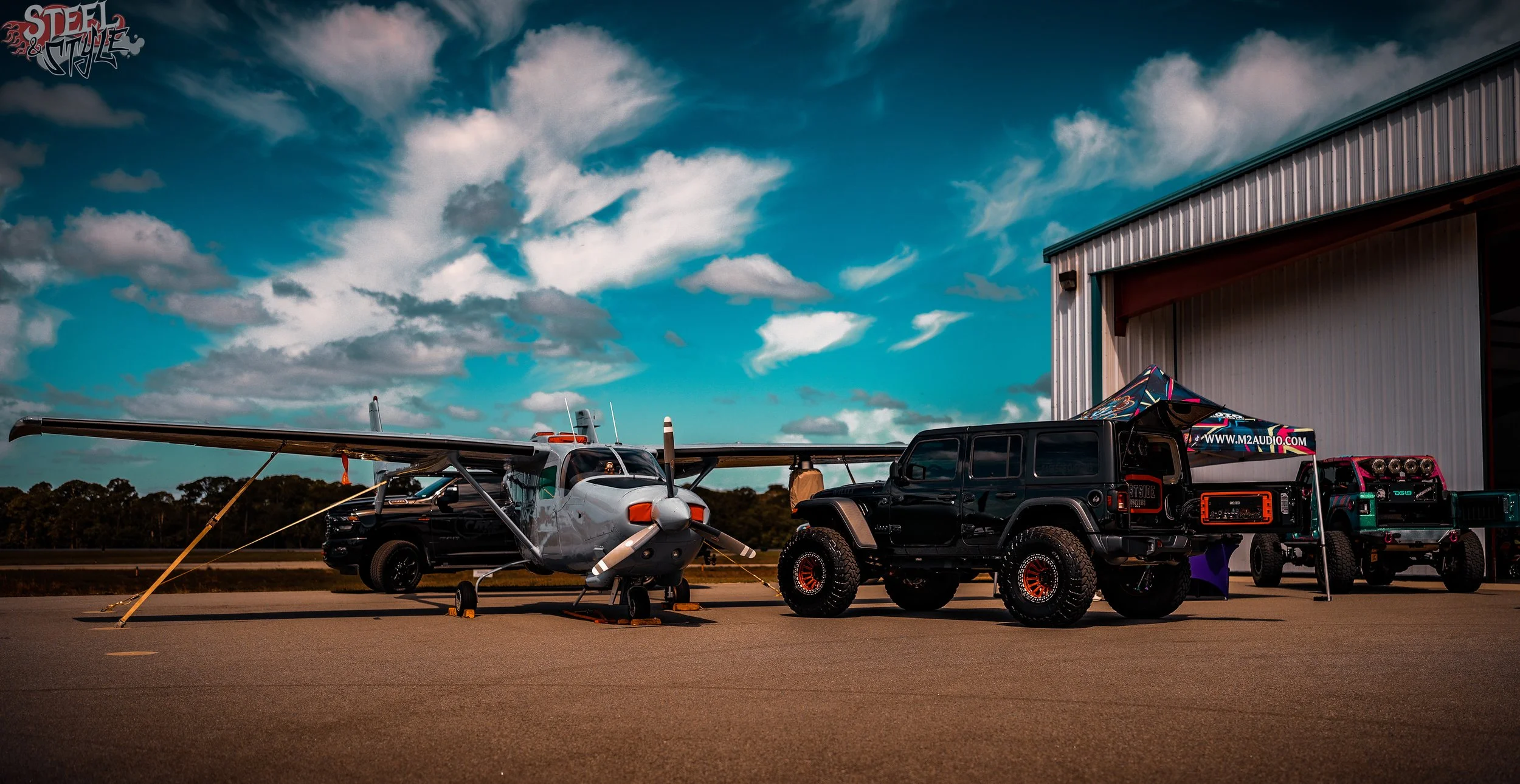 A small airplane parked on an airstrip next to a black Jeep and a tent with audio equipment, with a partly cloudy sky overhead.
