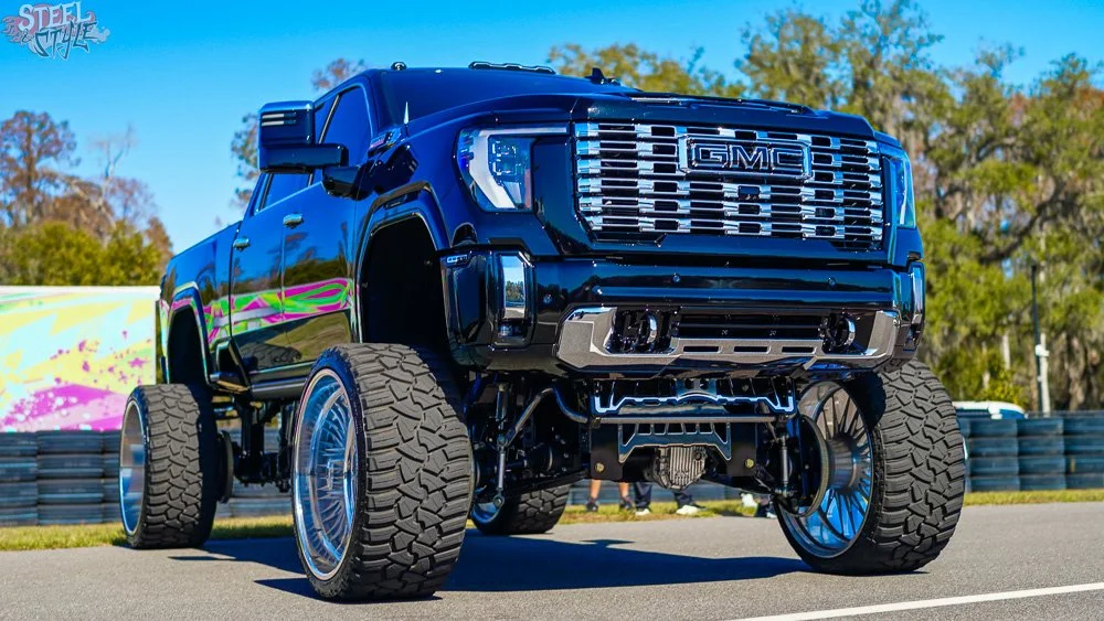A custom black GMC truck with large off-road tires, oversized rims, and lifted suspension, displayed outdoors at a car event.