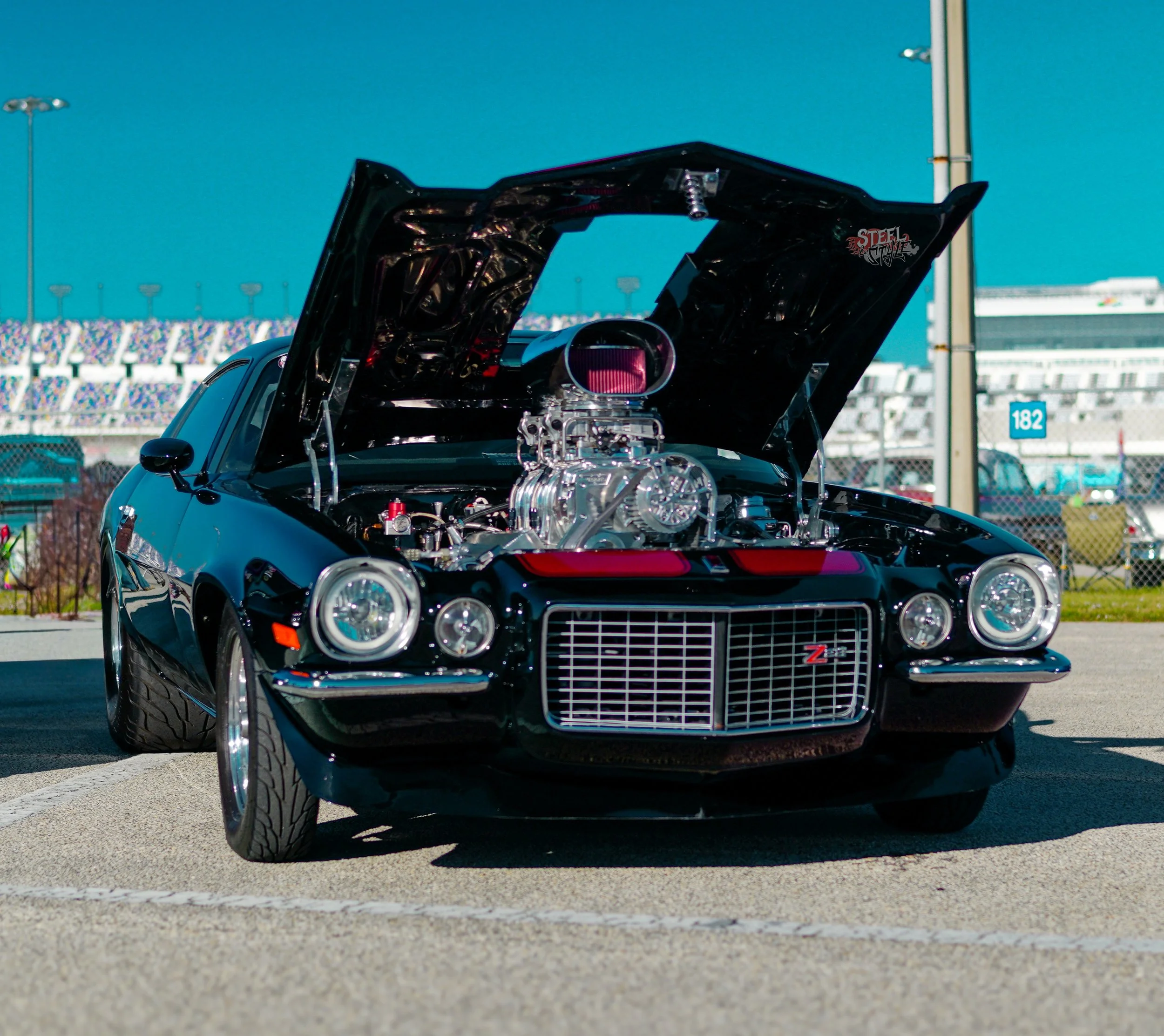 A black vintage Chevrolet Corvette ZR1 with an open hood displaying a polished engine, parked outdoors with a race track in the background.