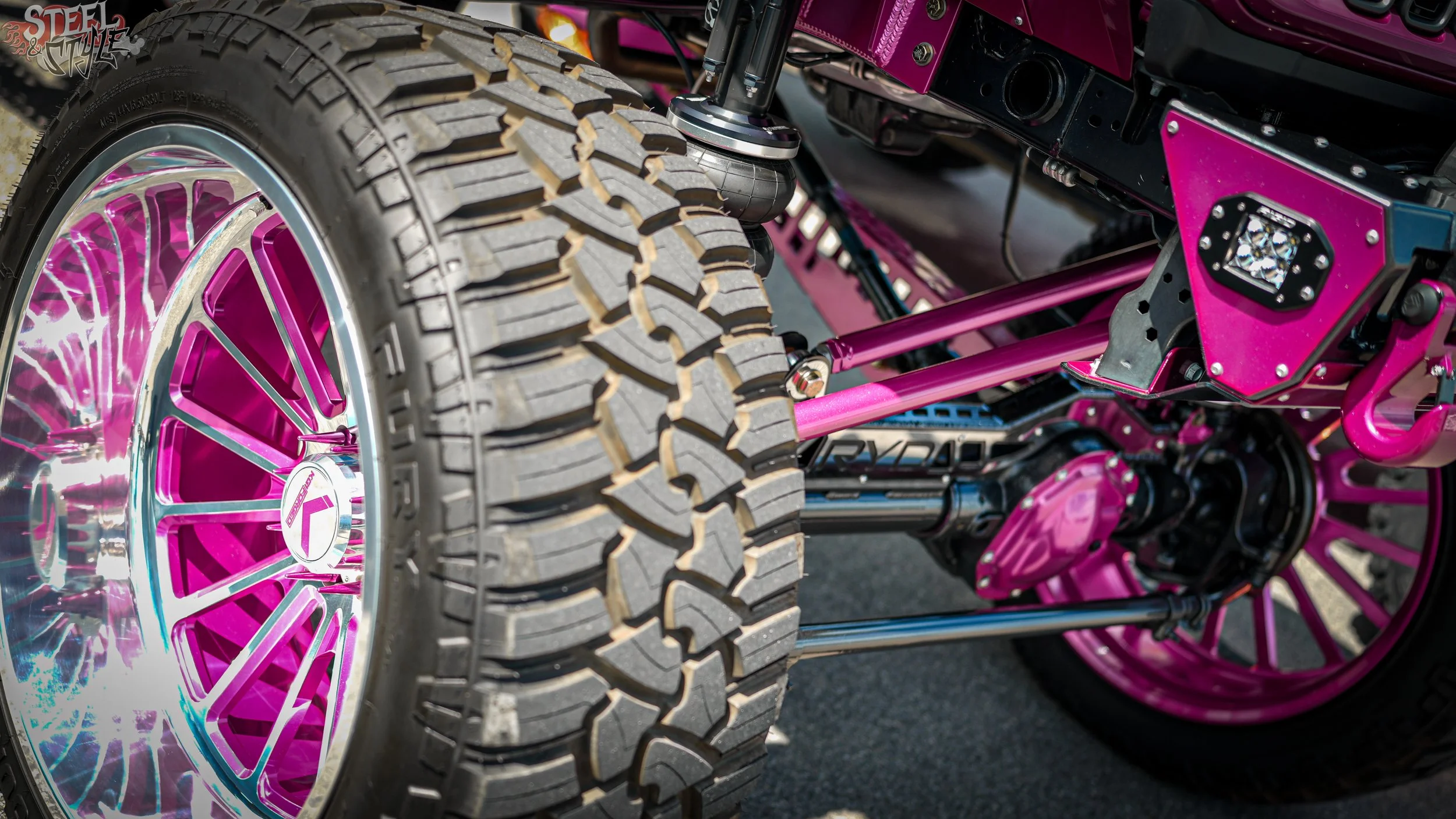 Close-up of a custom off-road vehicle with large knobby tires, pink rims, and chassis components in pink and black.