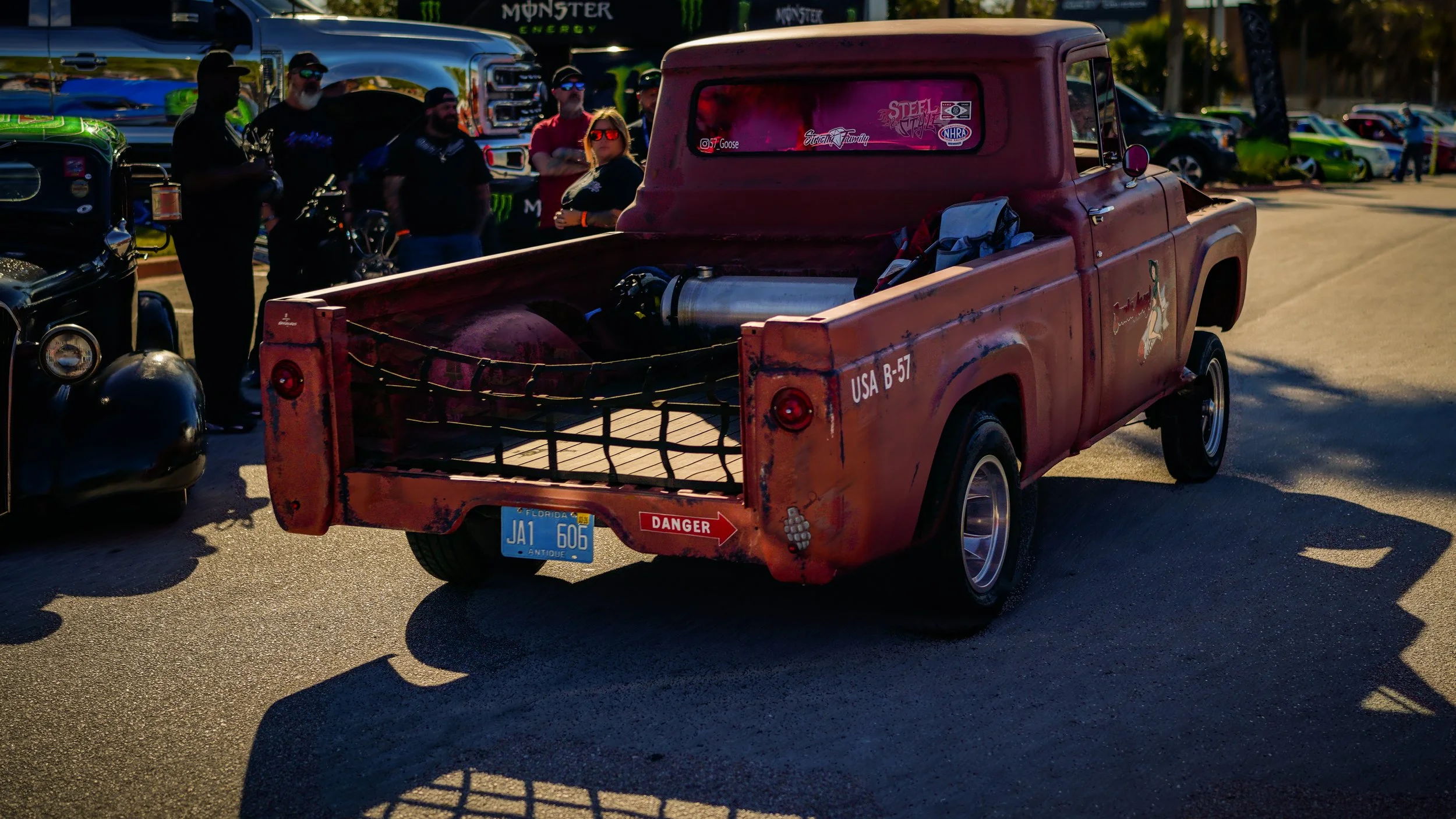 A vintage rust-colored pickup truck with a large fuel tank in the bed, parked among other classic cars and motorcycles at an outdoor car show, with people standing in the background near a Monster Energy truck.