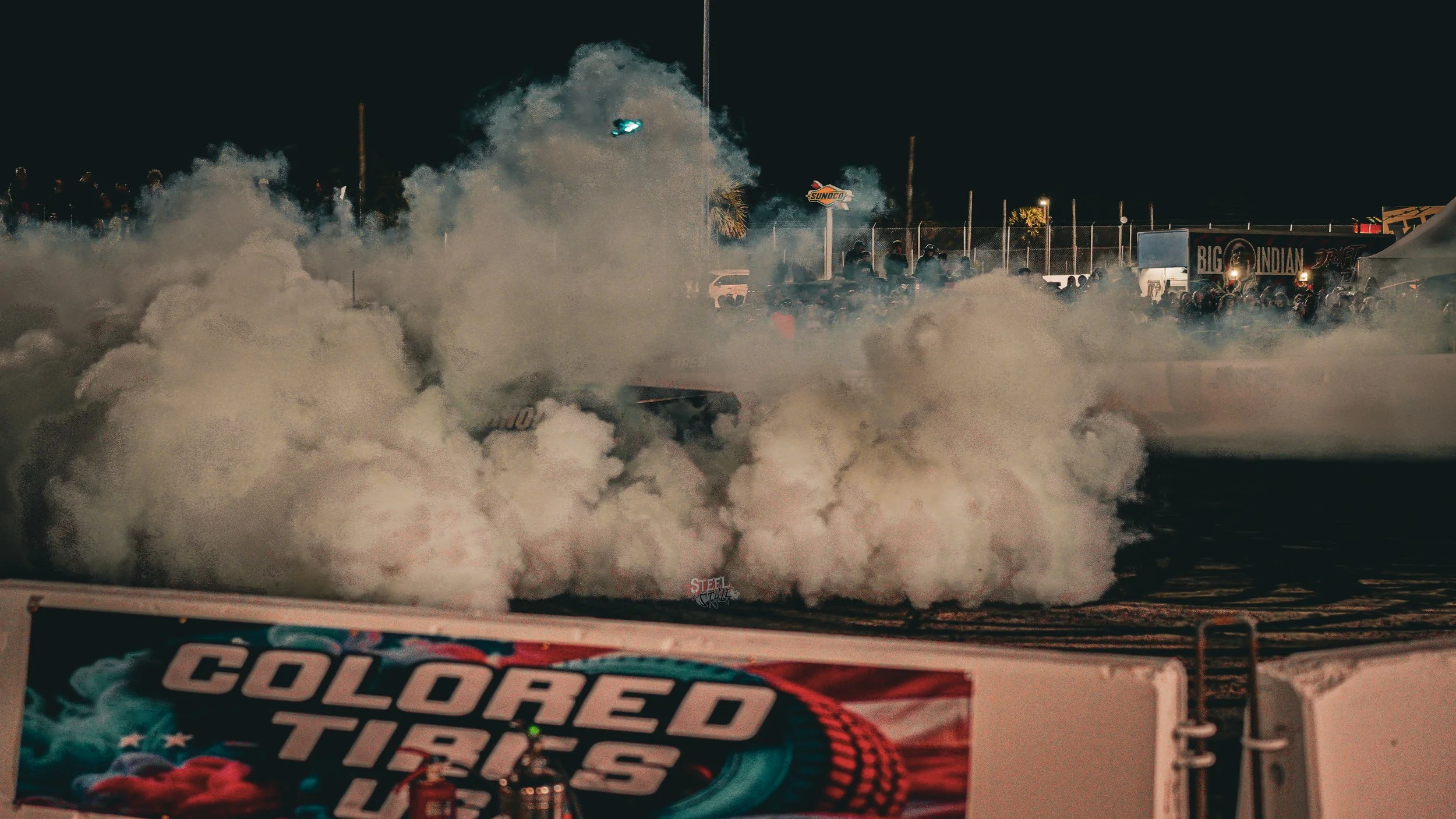 Race car performing a burnout on a racetrack at night, billowing smoke. Crowd watching in the background, with illuminated signs and tents.