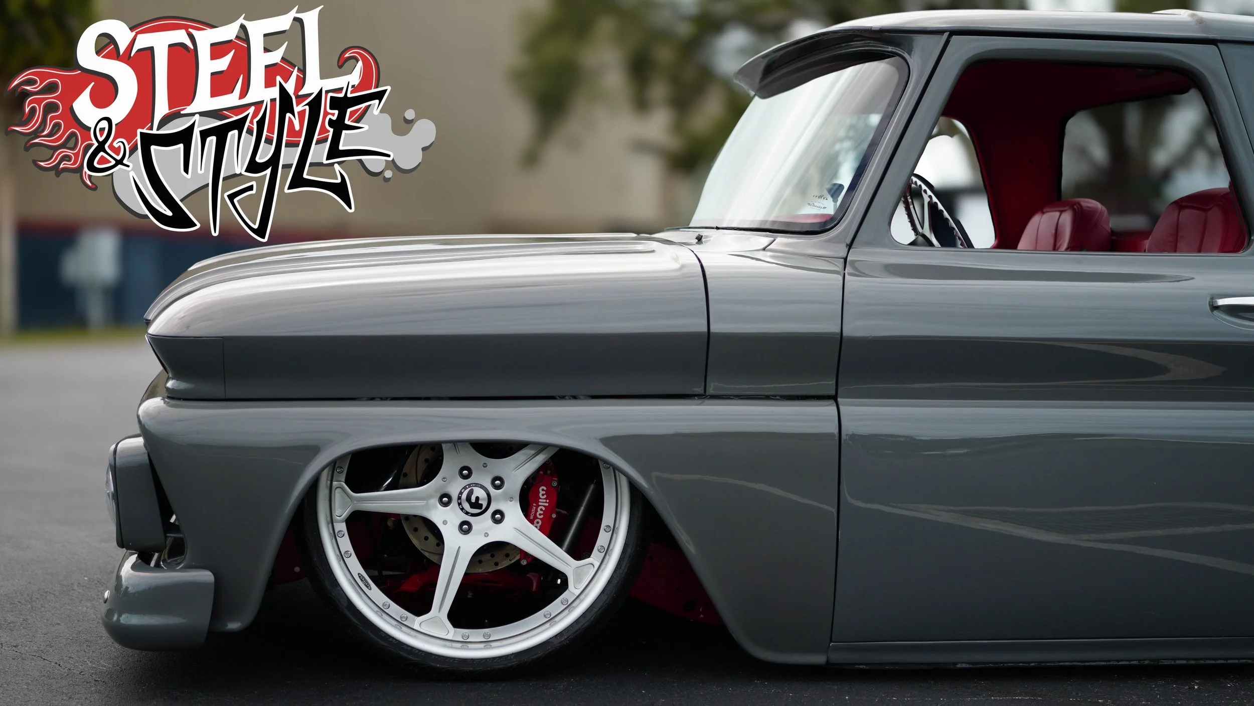 Close-up of a modified gray car with red interior, lowered stance, custom wheels, and racing brake calipers, with a 'Steel & Style' graffiti-style logo in the upper left corner.
