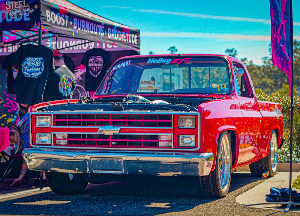 A red vintage Chevrolet pickup truck parked outdoors at a car show, with a display tent and merchandise in the background.