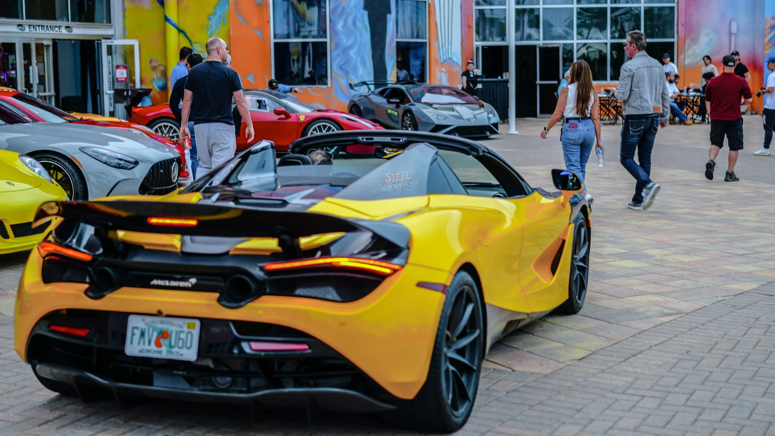Yellow convertible sports car parked at an outdoor exhibit, surrounded by other luxury cars and people walking and observing.
