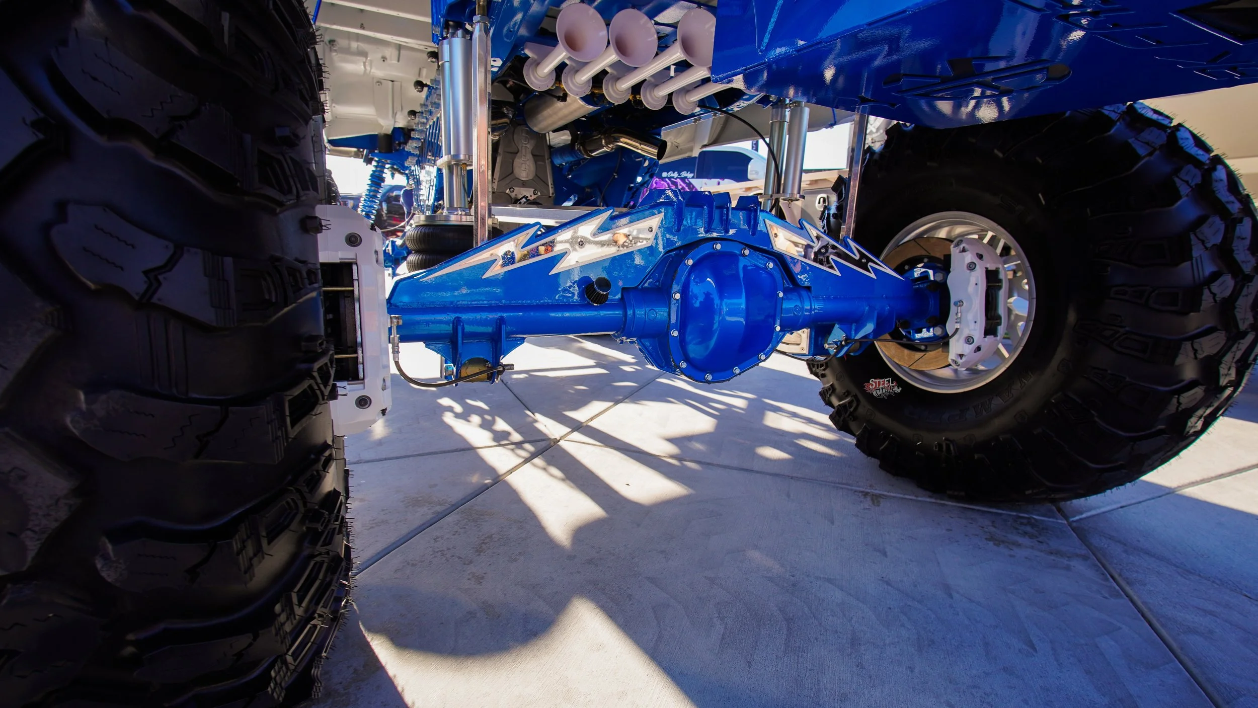 Close-up of a large off-road vehicle's suspension system and wheels, showing a bright blue axle, rugged tires, and parts of the suspension components.