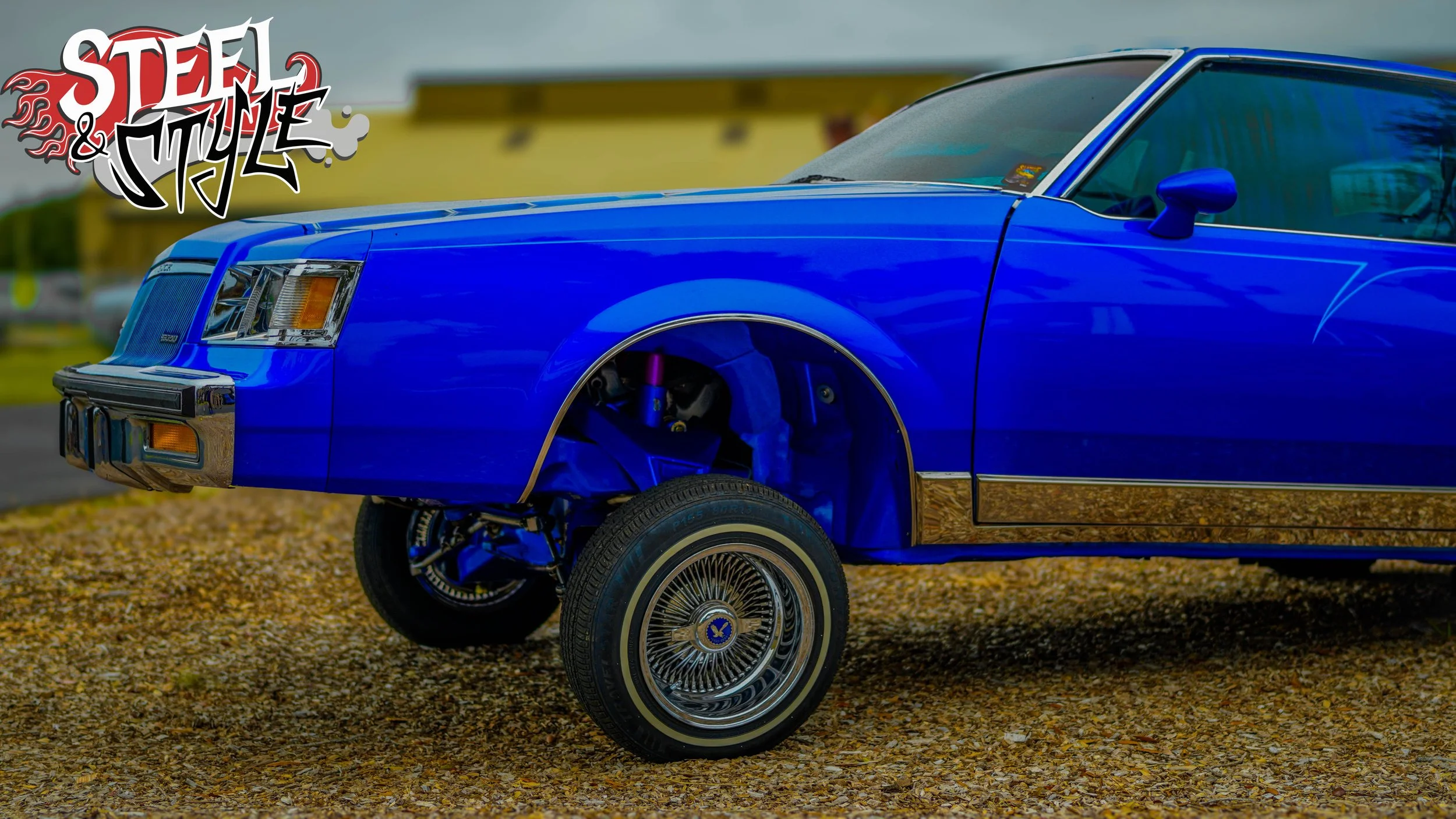 A bright blue vintage car with chrome accents, featuring wire-spoke wheels, parked on a gravel surface with a yellow building in the background. A graphic reading 'Steel & Style' is visible in the top left corner.