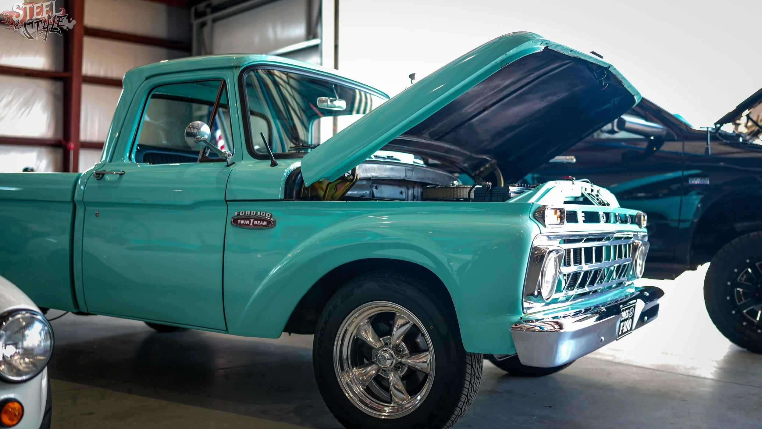 A vintage teal Ford F-100 pickup truck with its hood open, parked indoors alongside other vehicles.