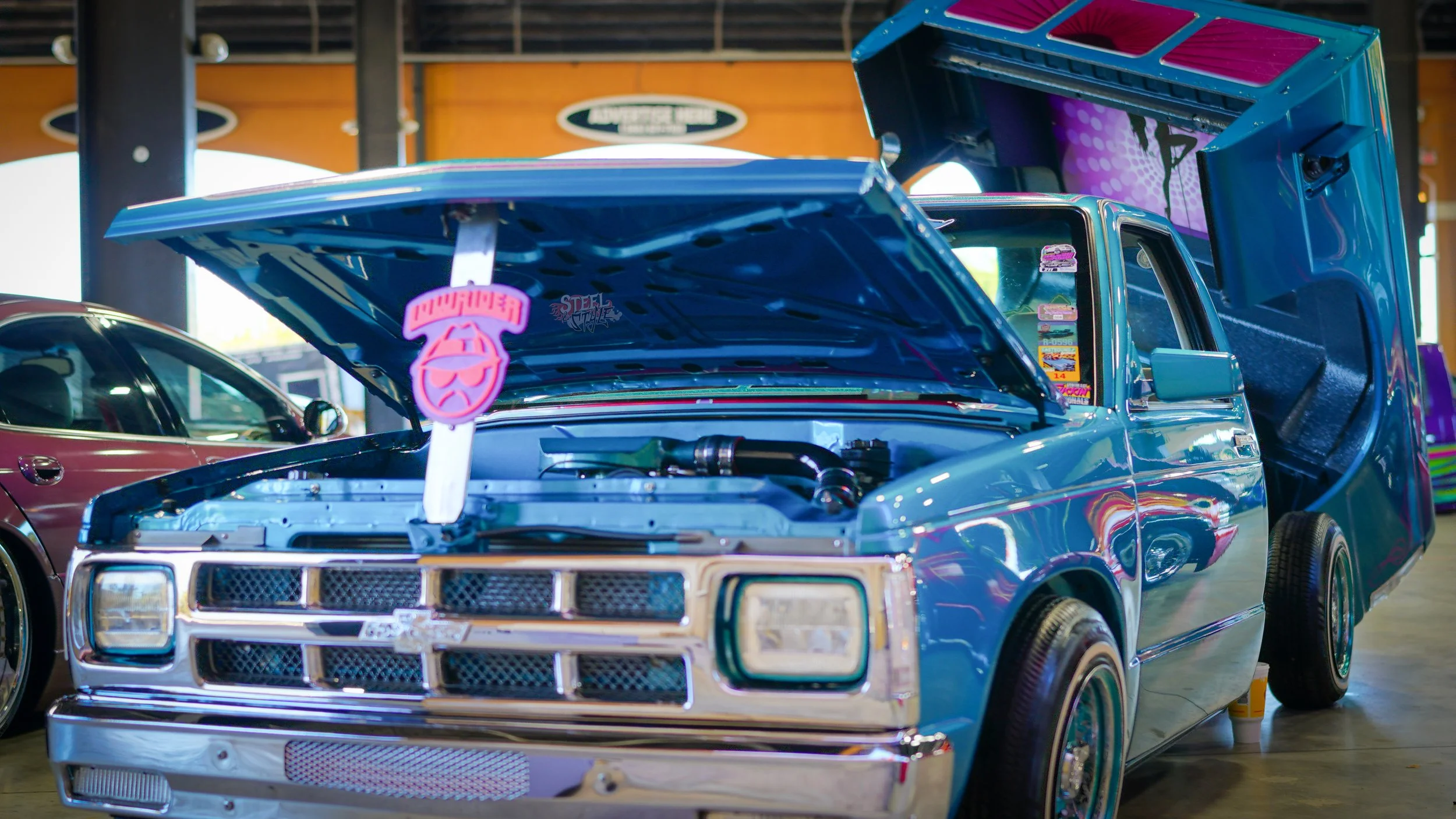 A vintage blue Chevrolet El Camino with the hood open and custom decals inside. The truck bed is lifted open, revealing its interior. The vehicle is displayed indoors, possibly at a car show.