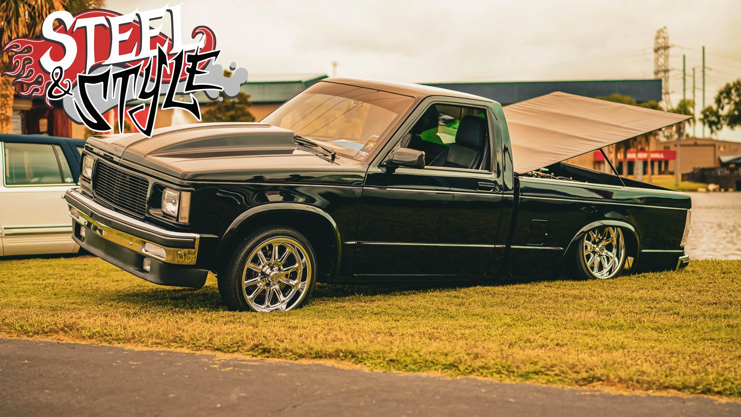 Black pickup truck with a tan sunshade covering the bed, parked on a grassy area near a body of water, with graffiti-style text saying 'Steel & Style' over the top left corner.