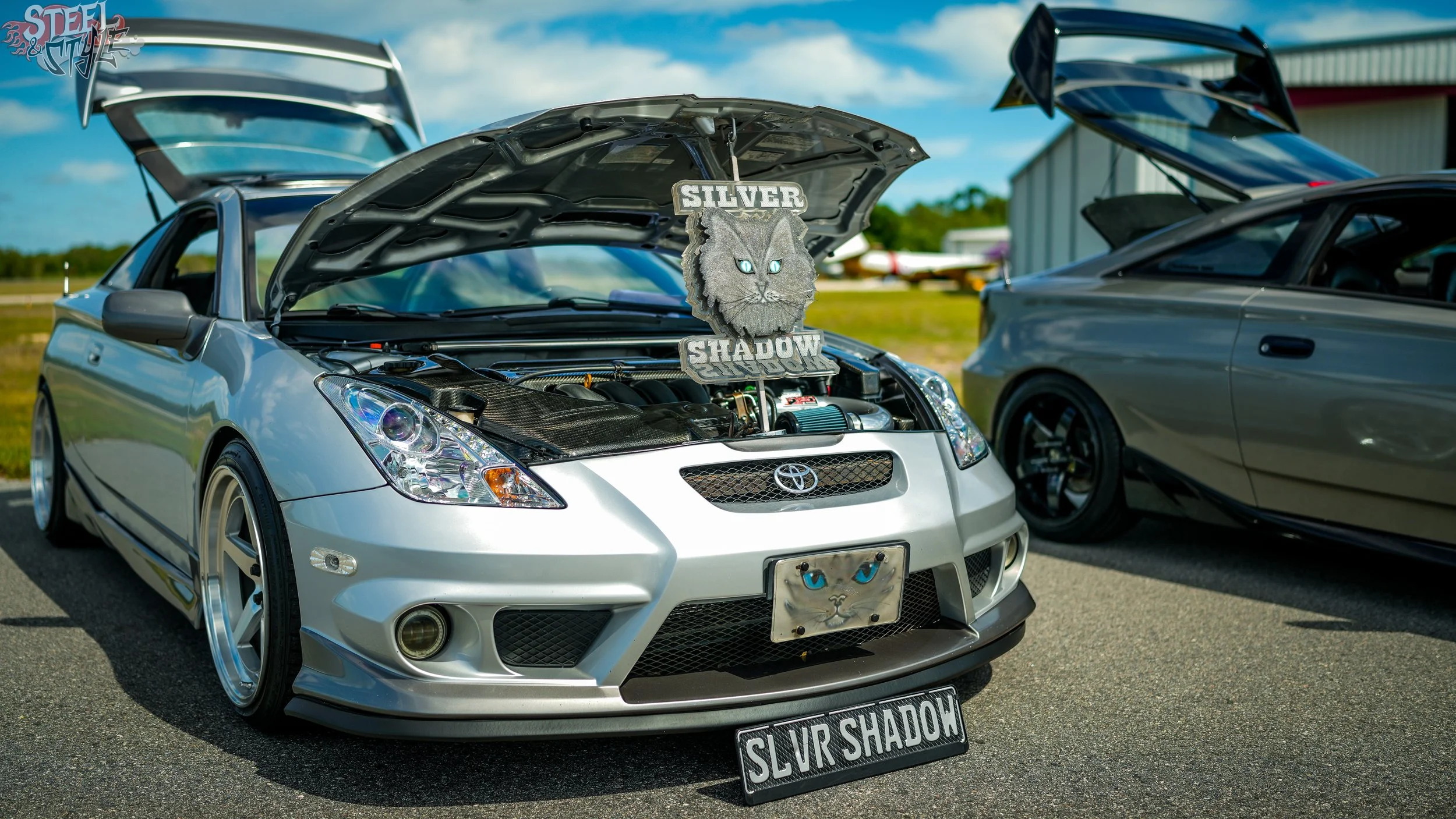 Silver Toyota car with hood open at a car show, featuring a decorative sign with a wolf's face and the words 'Silver Shadow' on the windshield. Other cars and a hangar are visible in the background on a sunny day.