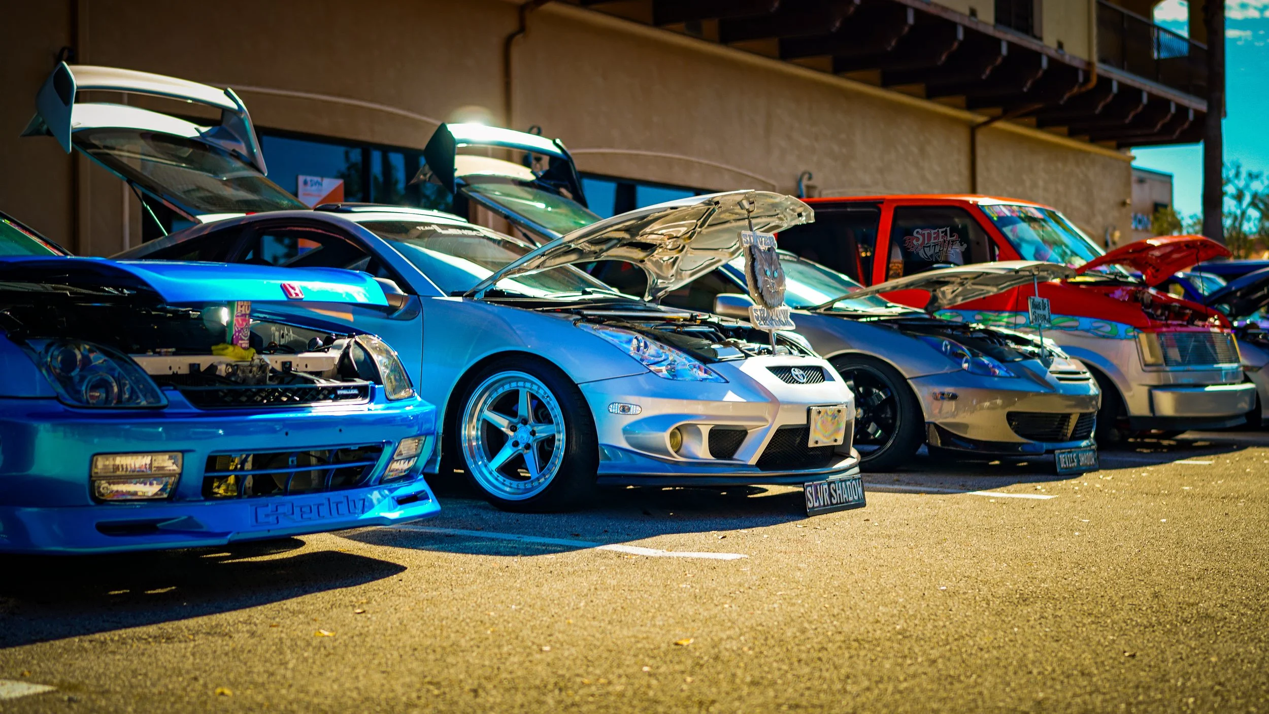 Line-up of various cars parked with hoods open at an outdoor car show during daytime.