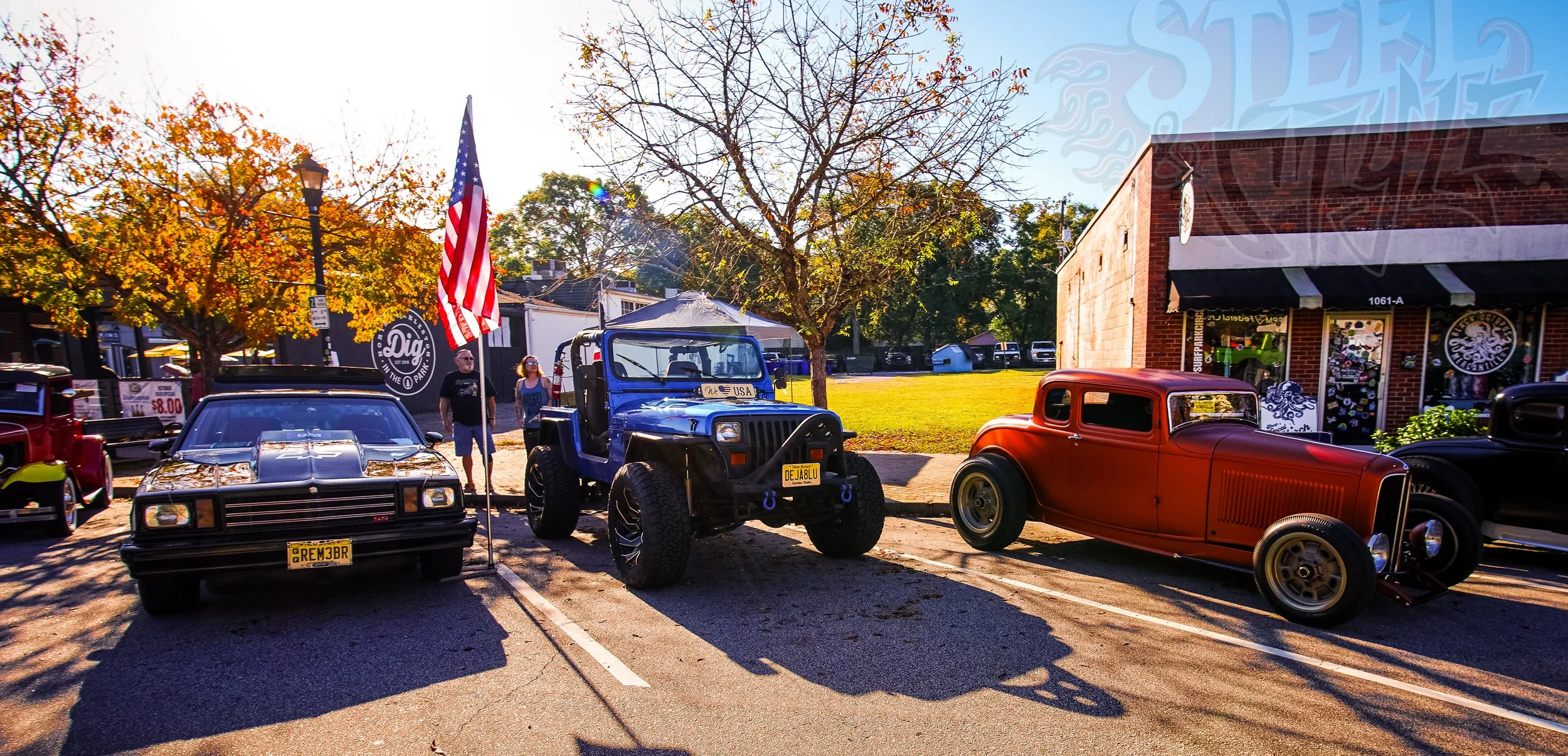 Three vintage cars parked in a row on a street during fall, with trees displaying orange and yellow leaves, and a building with shops in the background.