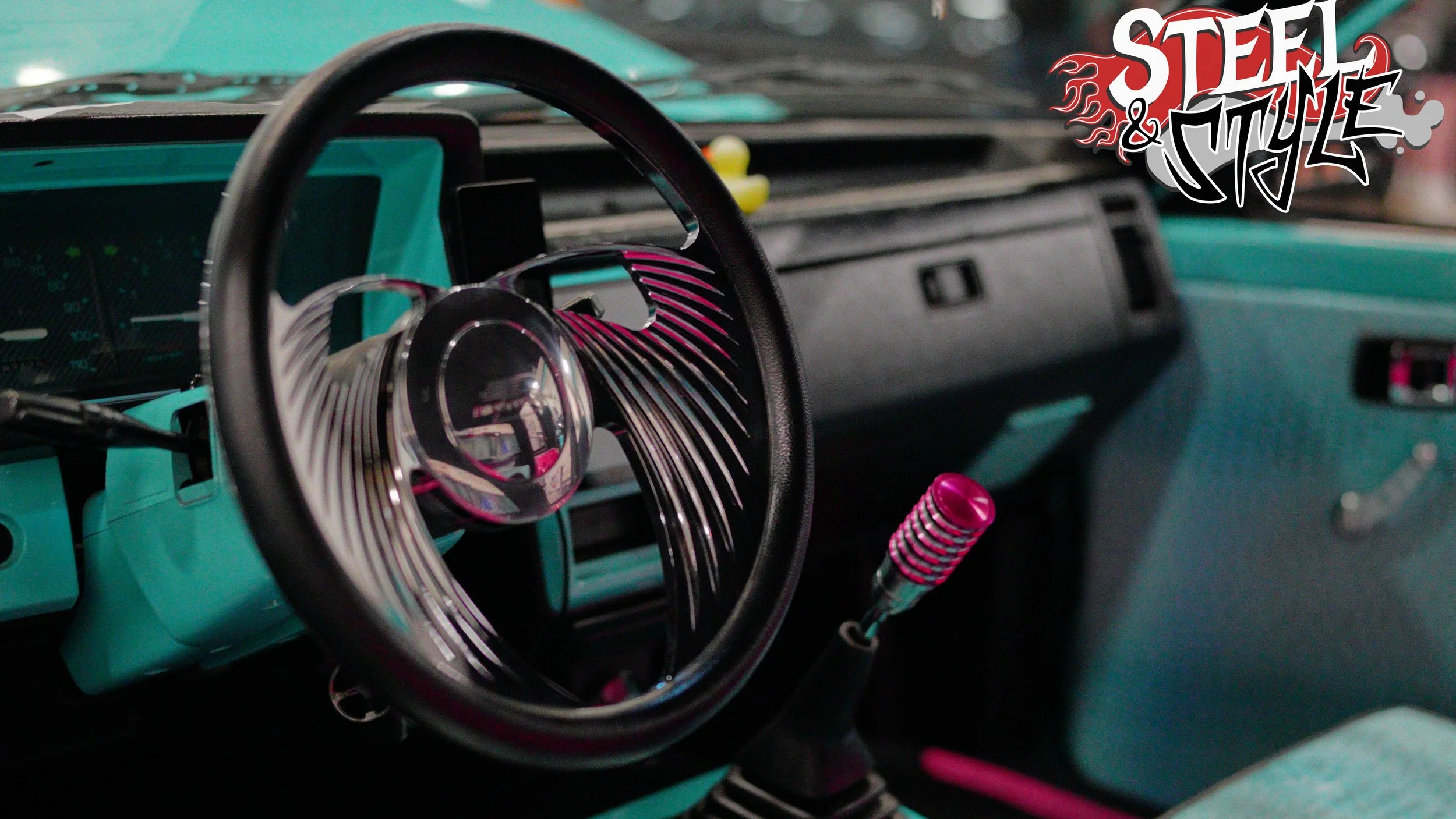 Interior of a vintage car with a turquoise dashboard, a black steering wheel with pink and black striped details, and a custom pink gear shift knob. The upper right corner has a 'Steel & Style' logo.
