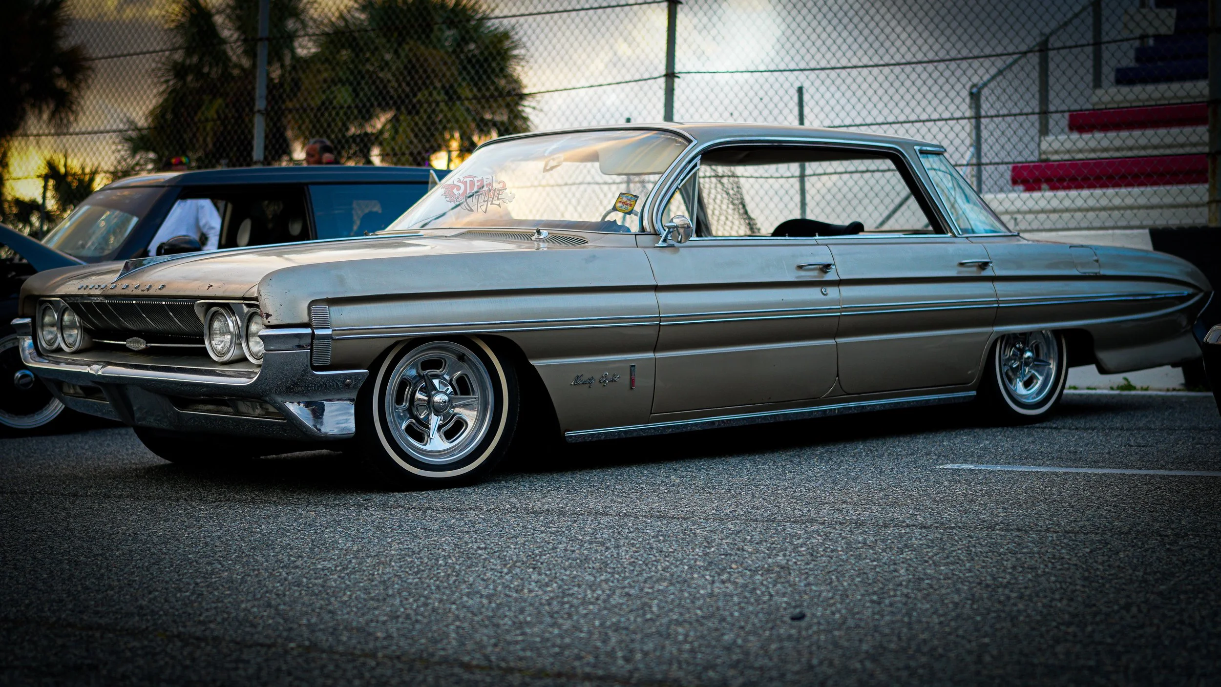 A vintage silver Mercury Monterey parked on a street during dusk, with a chain-link fence and trees in the background.