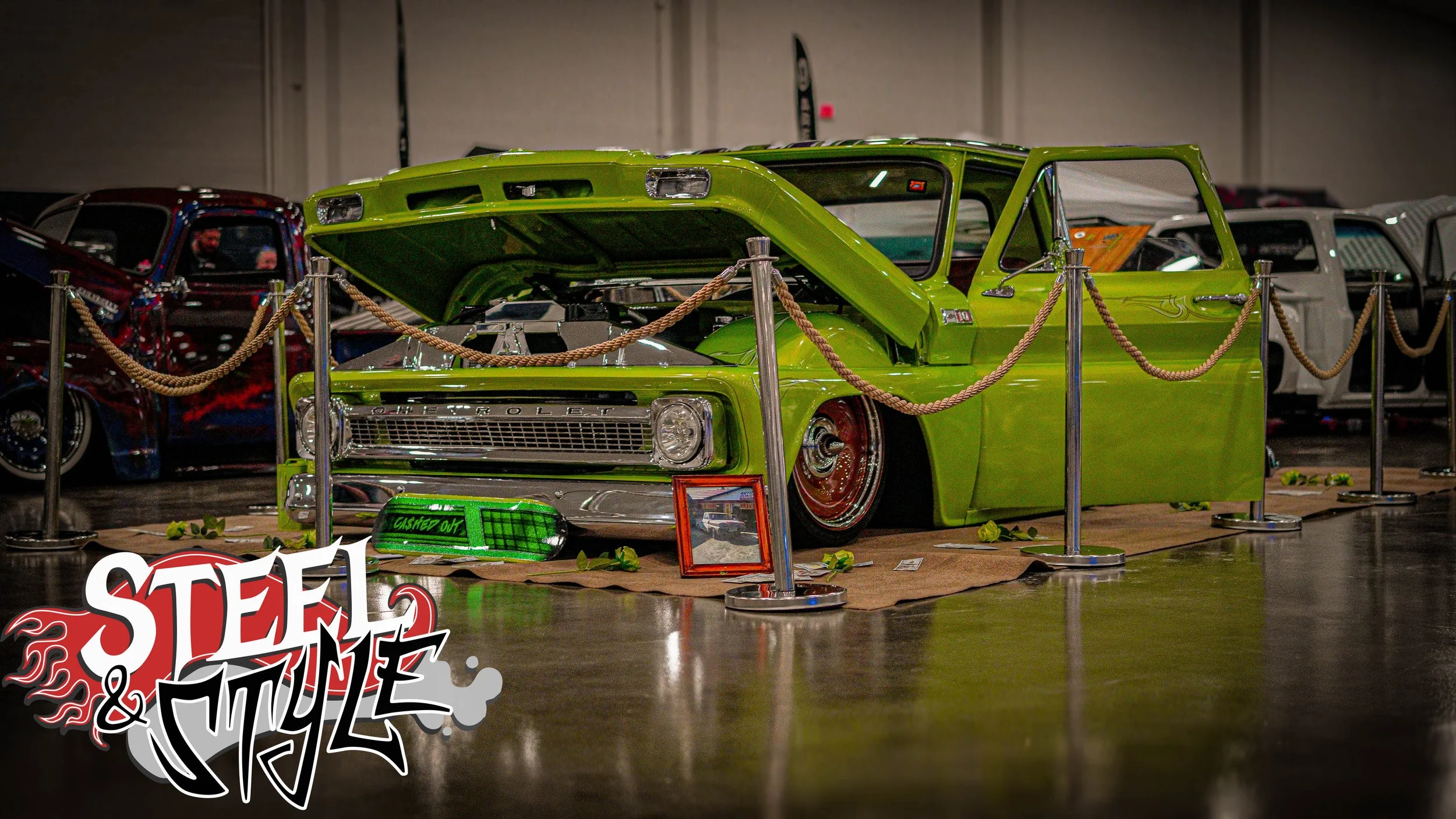 A customized classic green Chevrolet car on display at an indoor show, with its hood open and surrounded by ropes. There is a logo at the bottom left that says 'Steel & Style' with a flaming design.