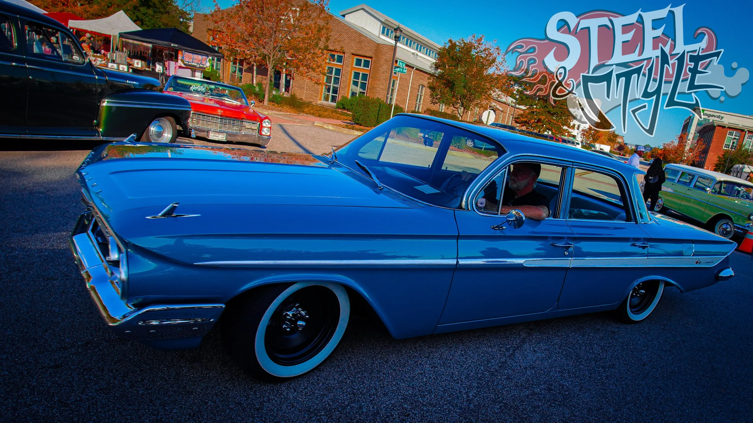 A vintage blue car is parked at a car show with other classic cars in the background and a sign reading 'Steel & Style' in the upper right corner.