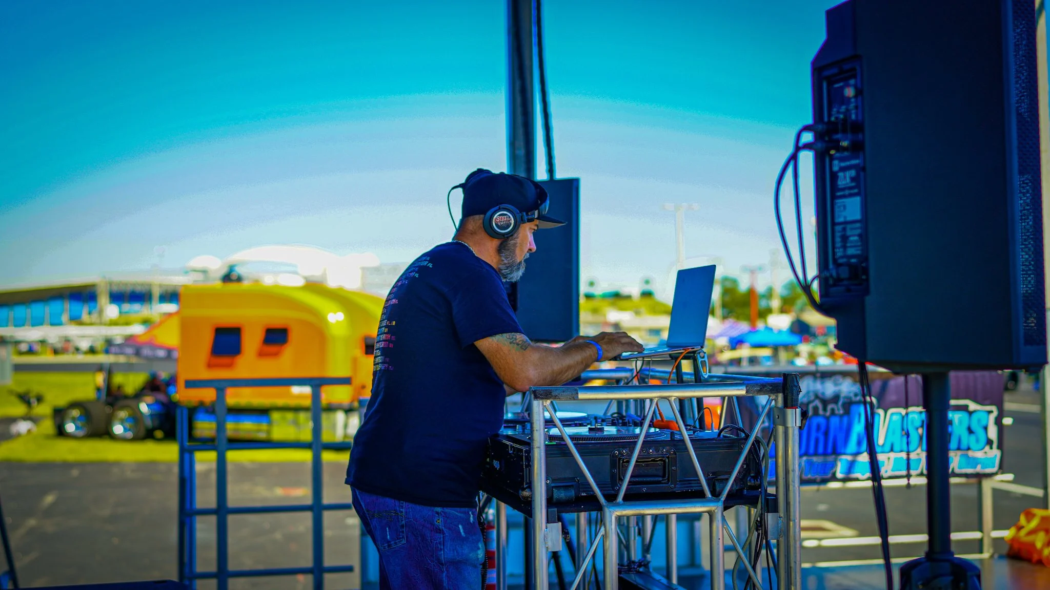 A DJ with headphones working on a laptop at an outdoor event during daytime, with a yellow food truck and tents in the background.