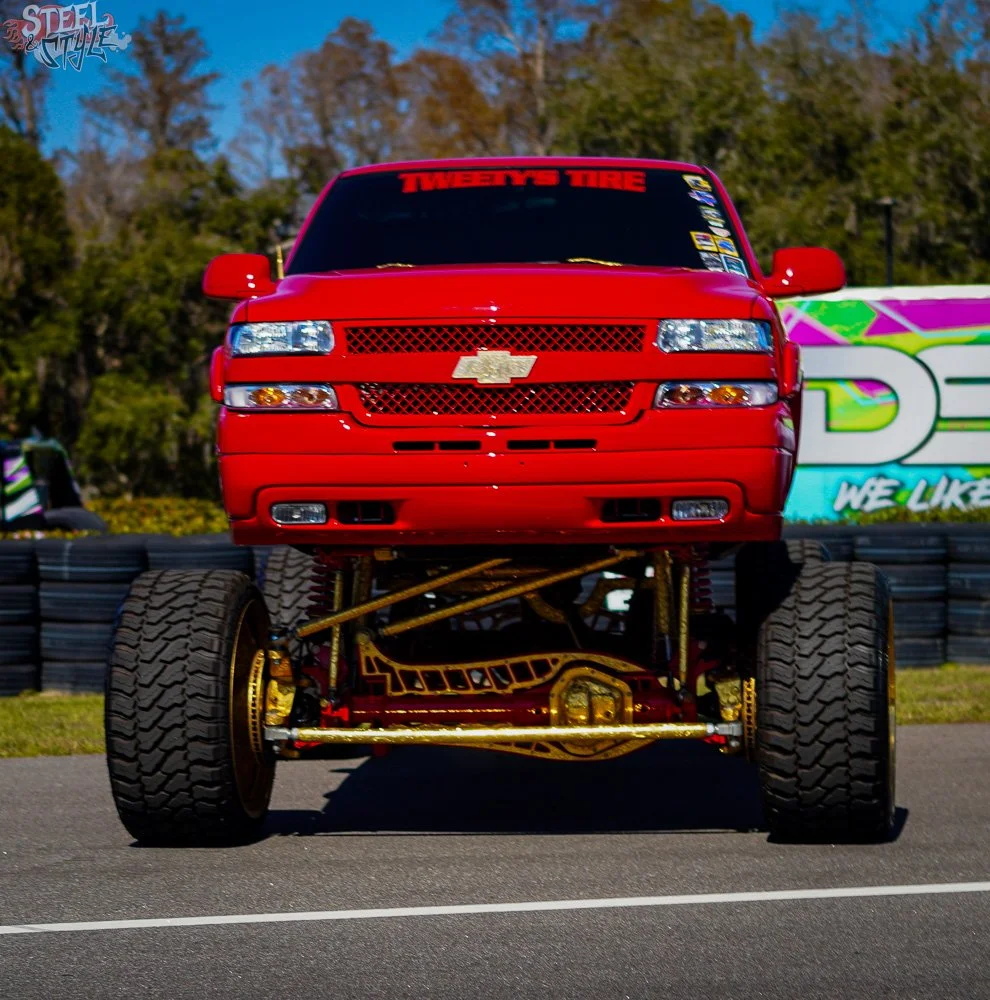 A red Chevrolet truck with oversized wheels and suspension, parked on a racetrack with a colorful advertisement banner in the background