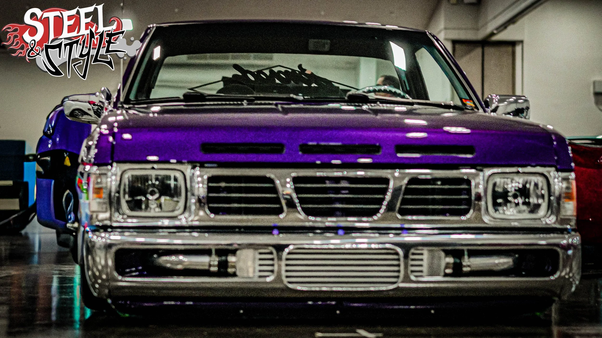 A vintage pickup truck with a vibrant purple and chrome paint job, parked indoors at a car show.