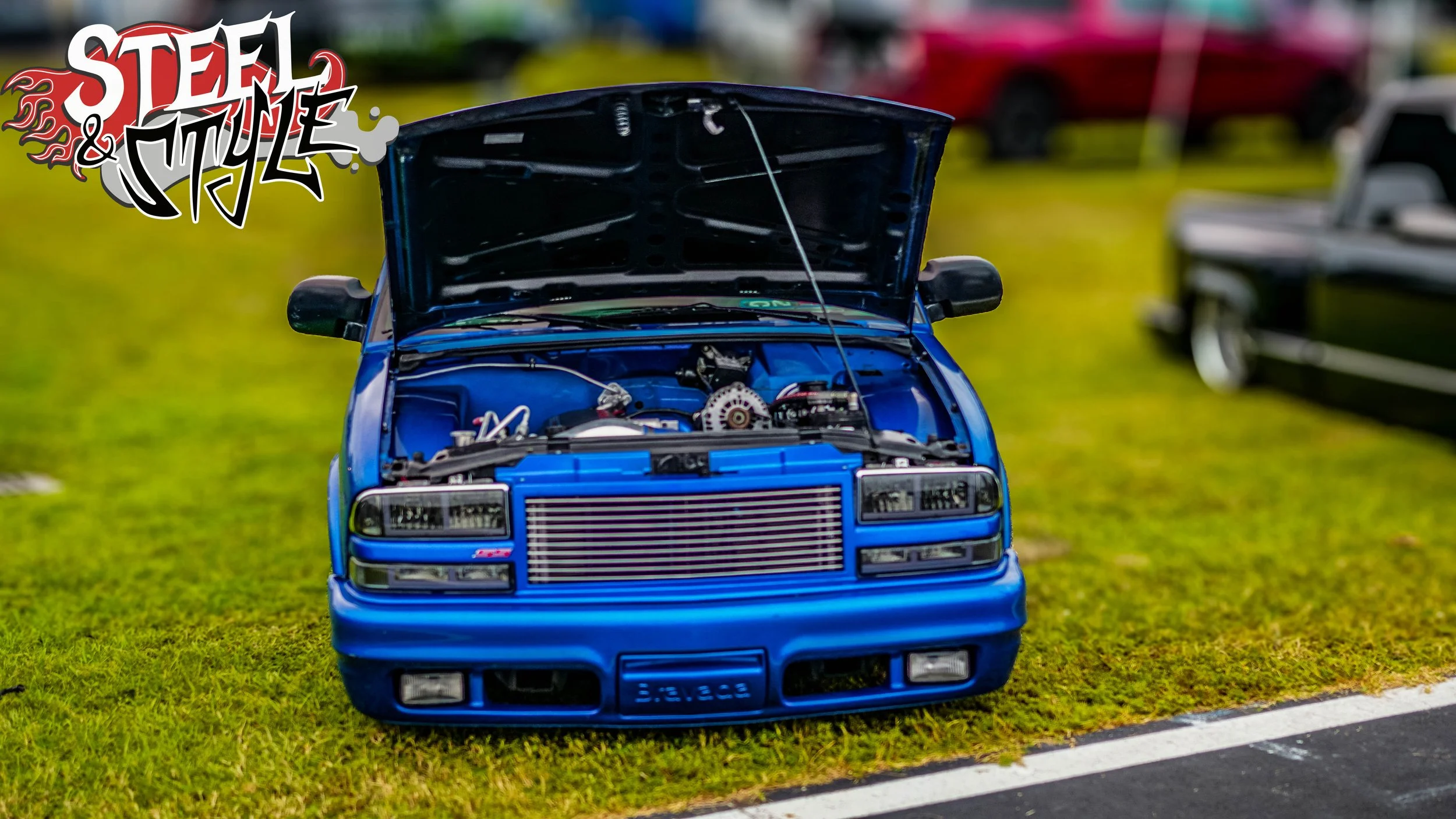 A blue car with its hood open, showing a modified engine and intercooler, parked on grass beside a road during daylight at a car show.