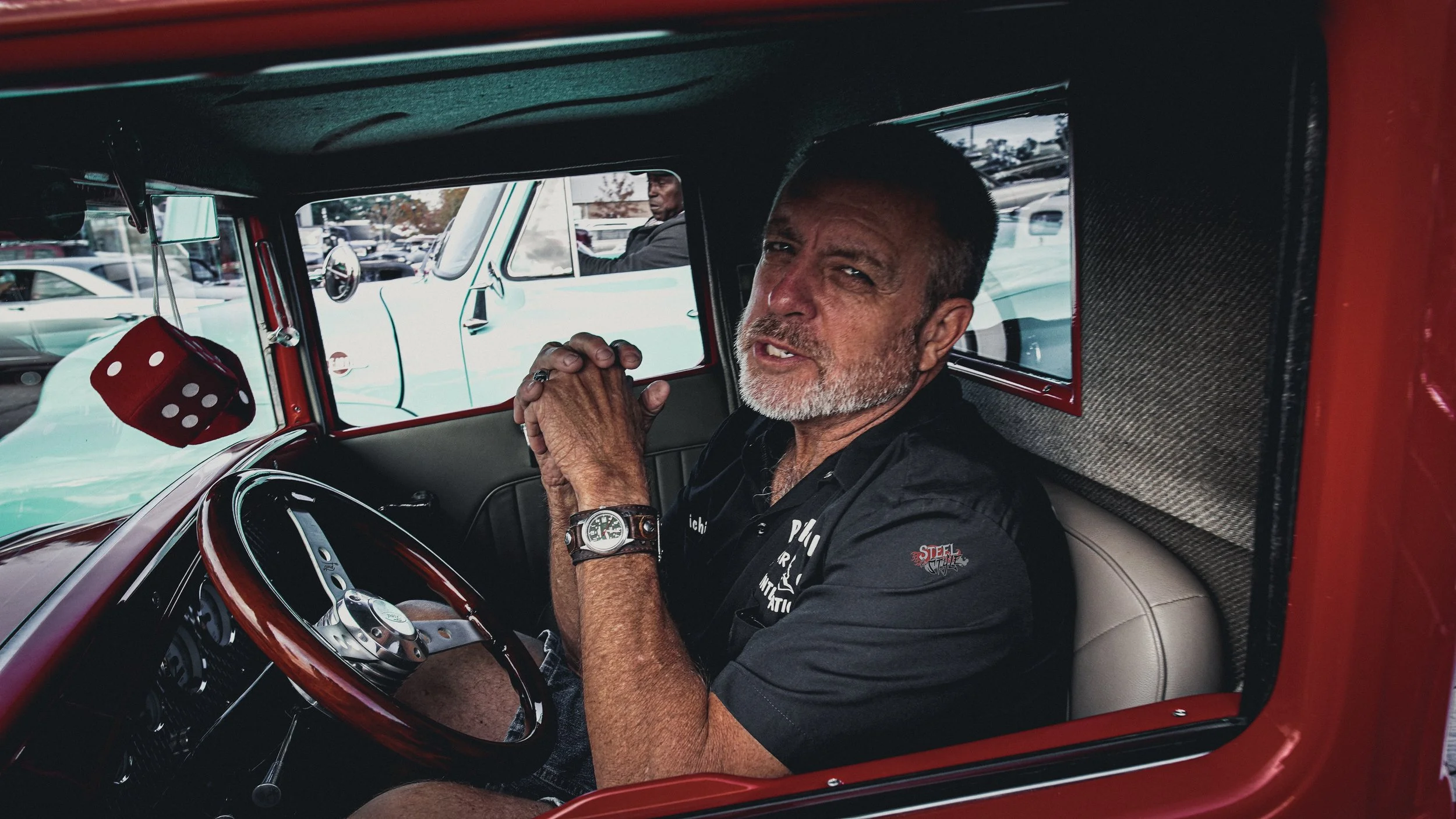 A man sitting inside a vintage red car, looking at the camera, with a parking lot and other vehicles visible outside the window.