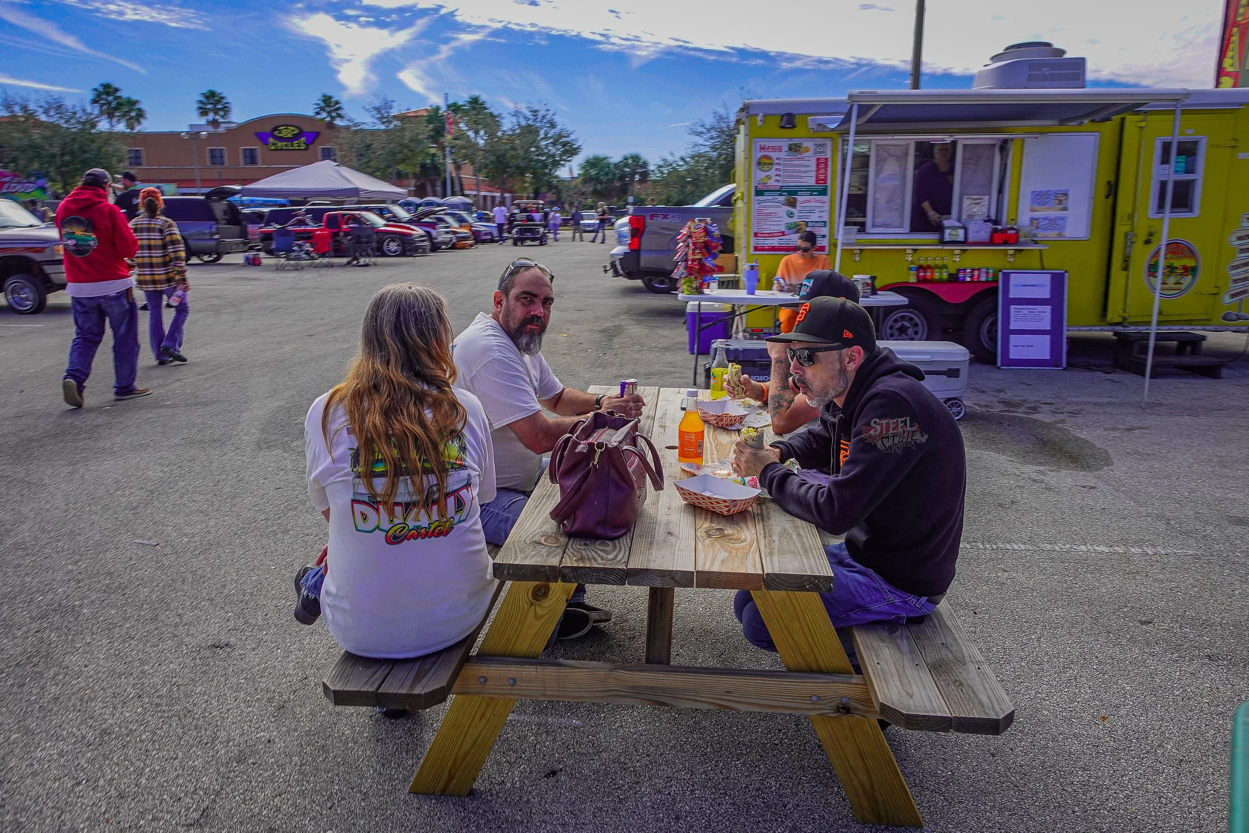People sitting at a picnic table at an outdoor food festival with food trucks and parked cars in the background.
