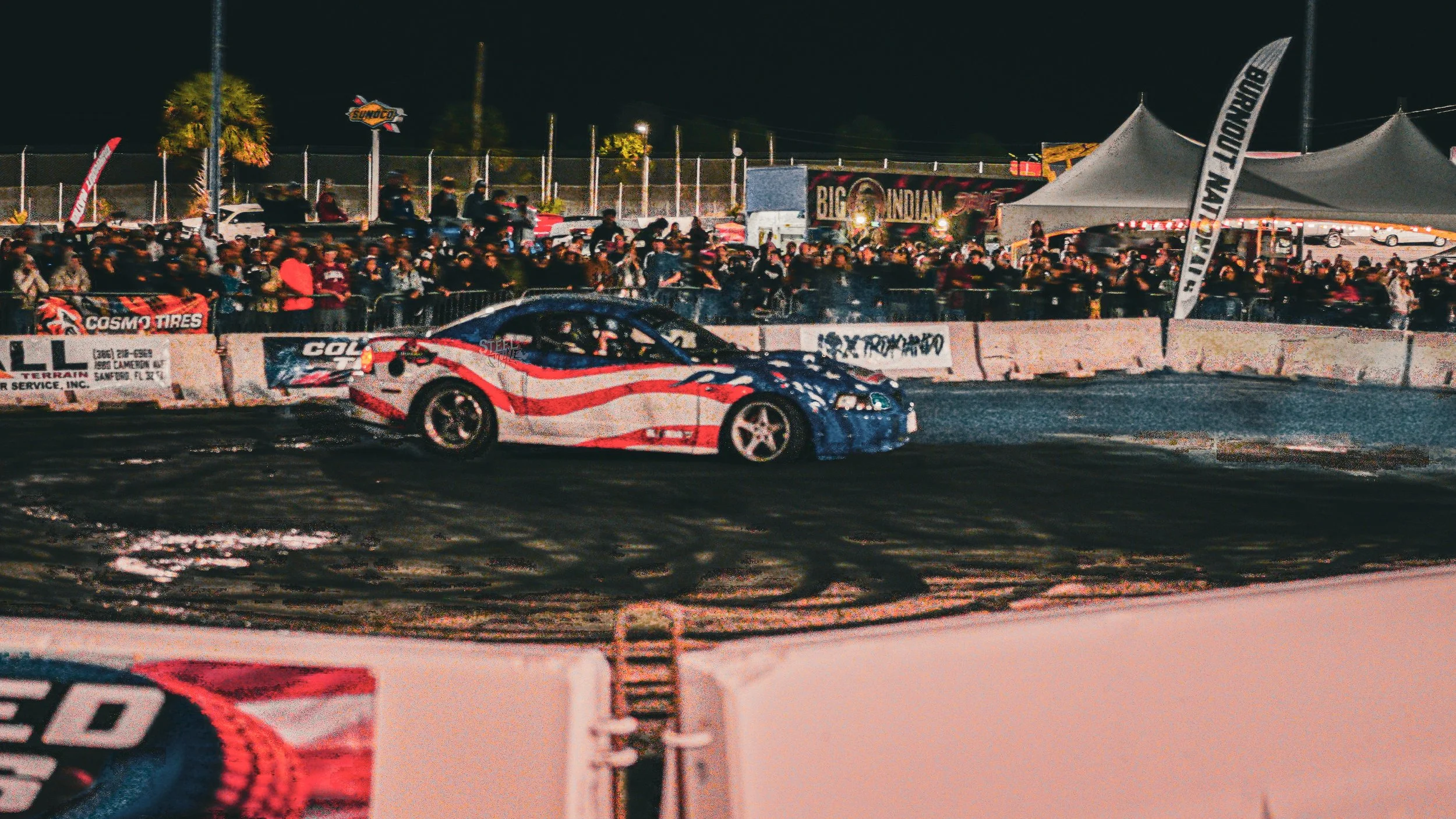 Nighttime scene of a race car drifting around a track, with a large crowd of spectators behind barriers and tents, illuminated by bright lights.
