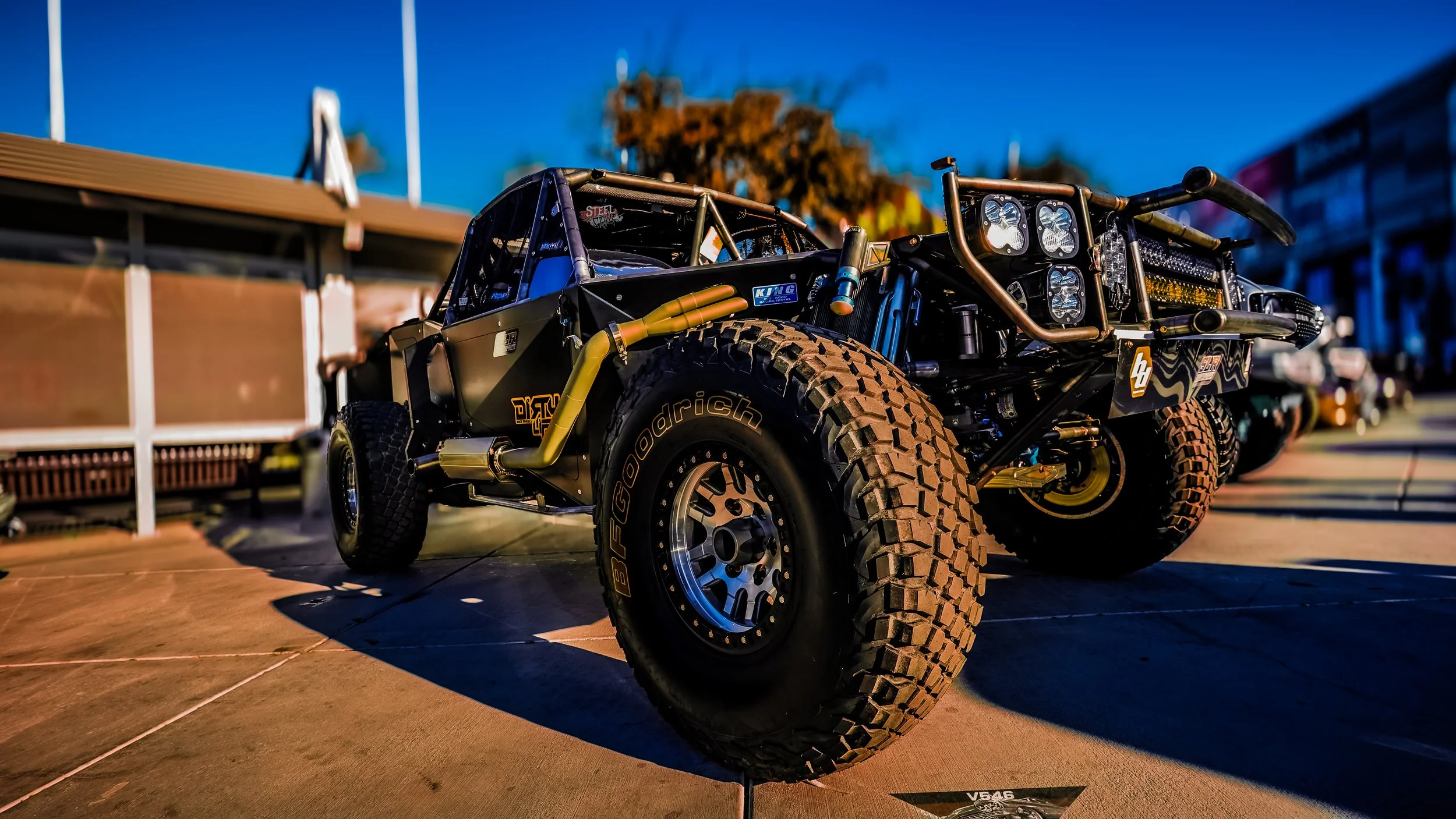 Off-road race truck parked on concrete surface, with large tires, custom suspension, and multiple lights, during sunset.