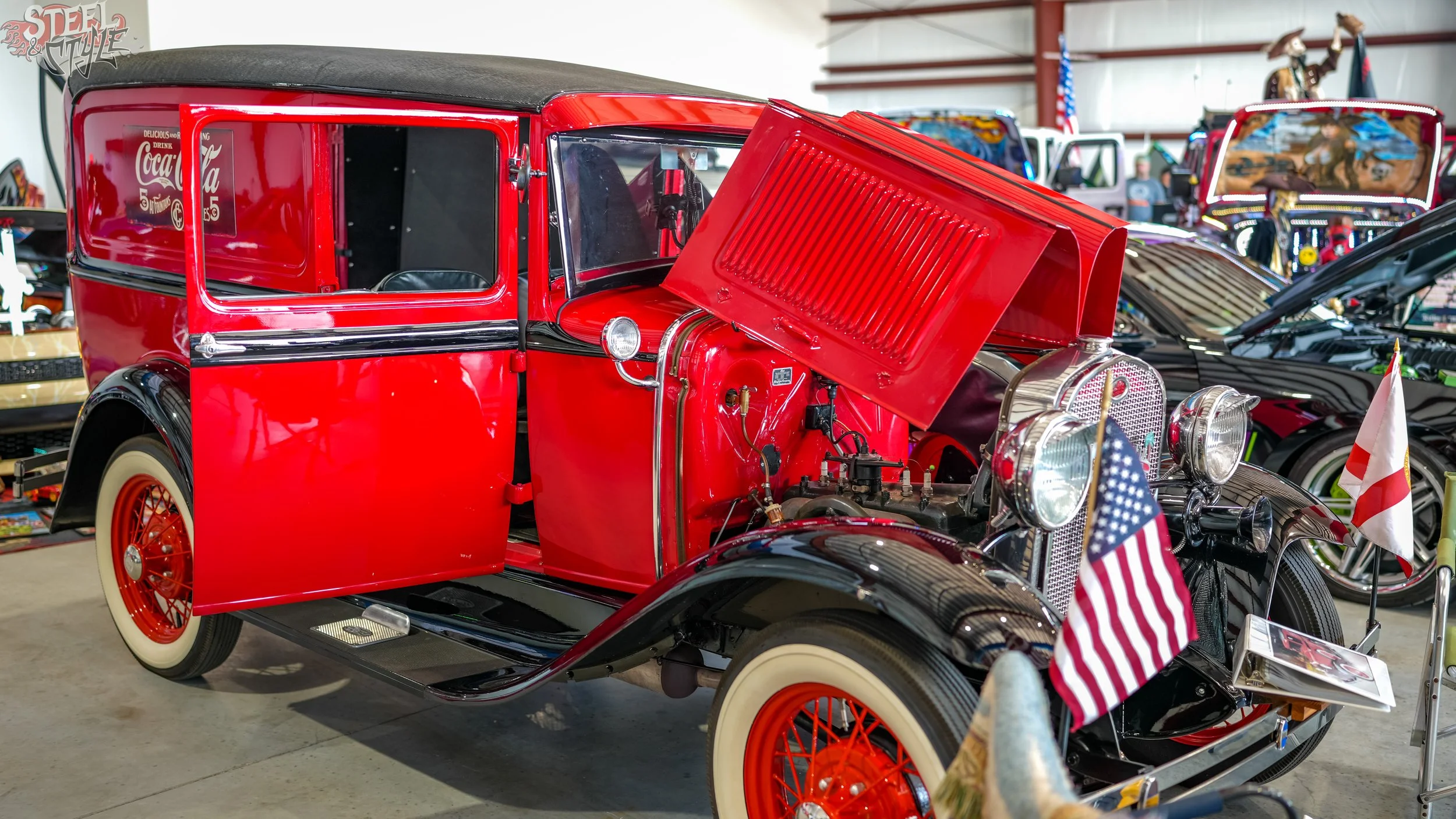 A vintage red and black automobile with its hood open on display in an indoor exhibit, with American flags and other cars in the background.