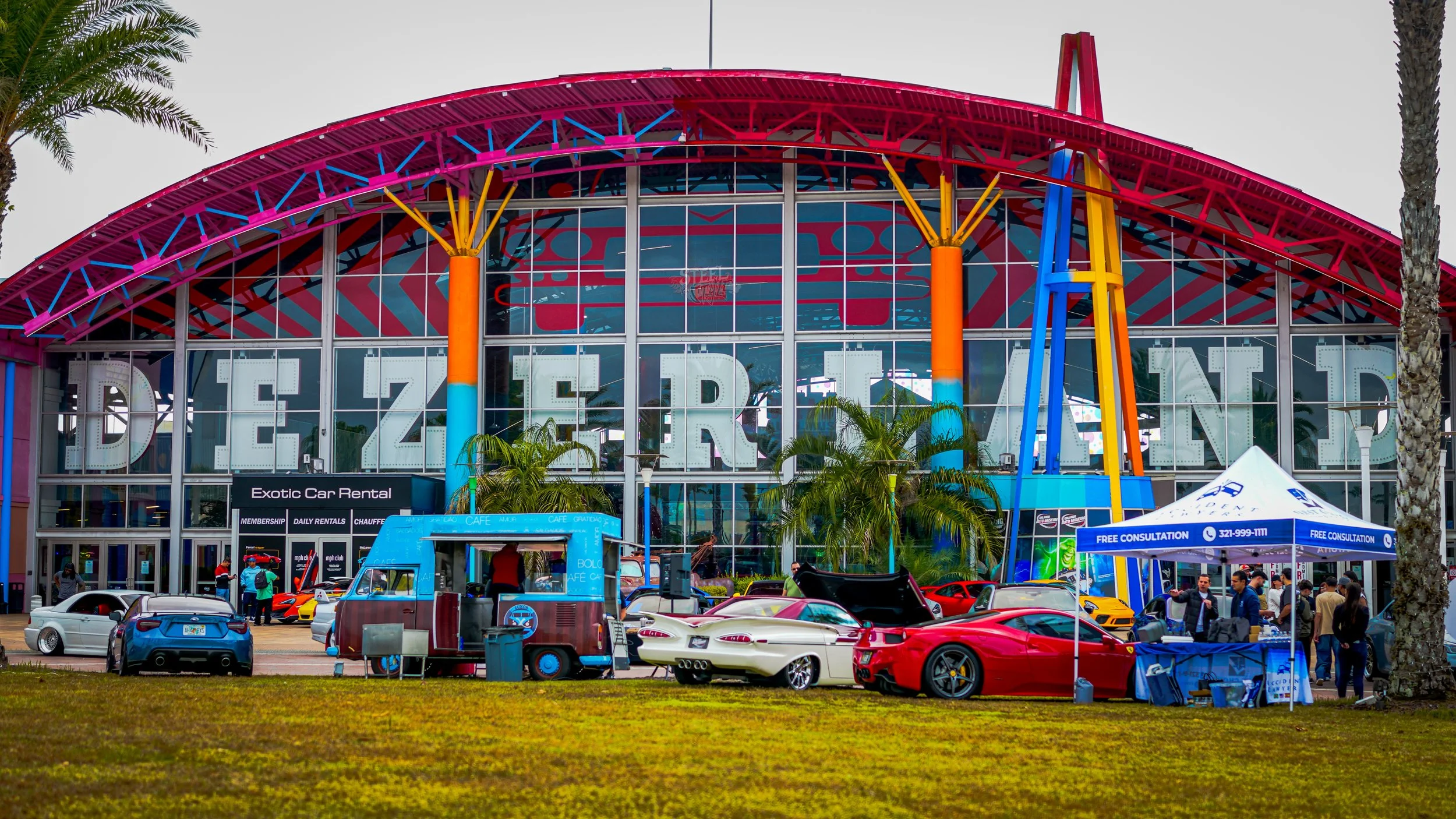 Colorful building with large windows, palm trees, and several luxury cars parked in front, at an outdoor car event.