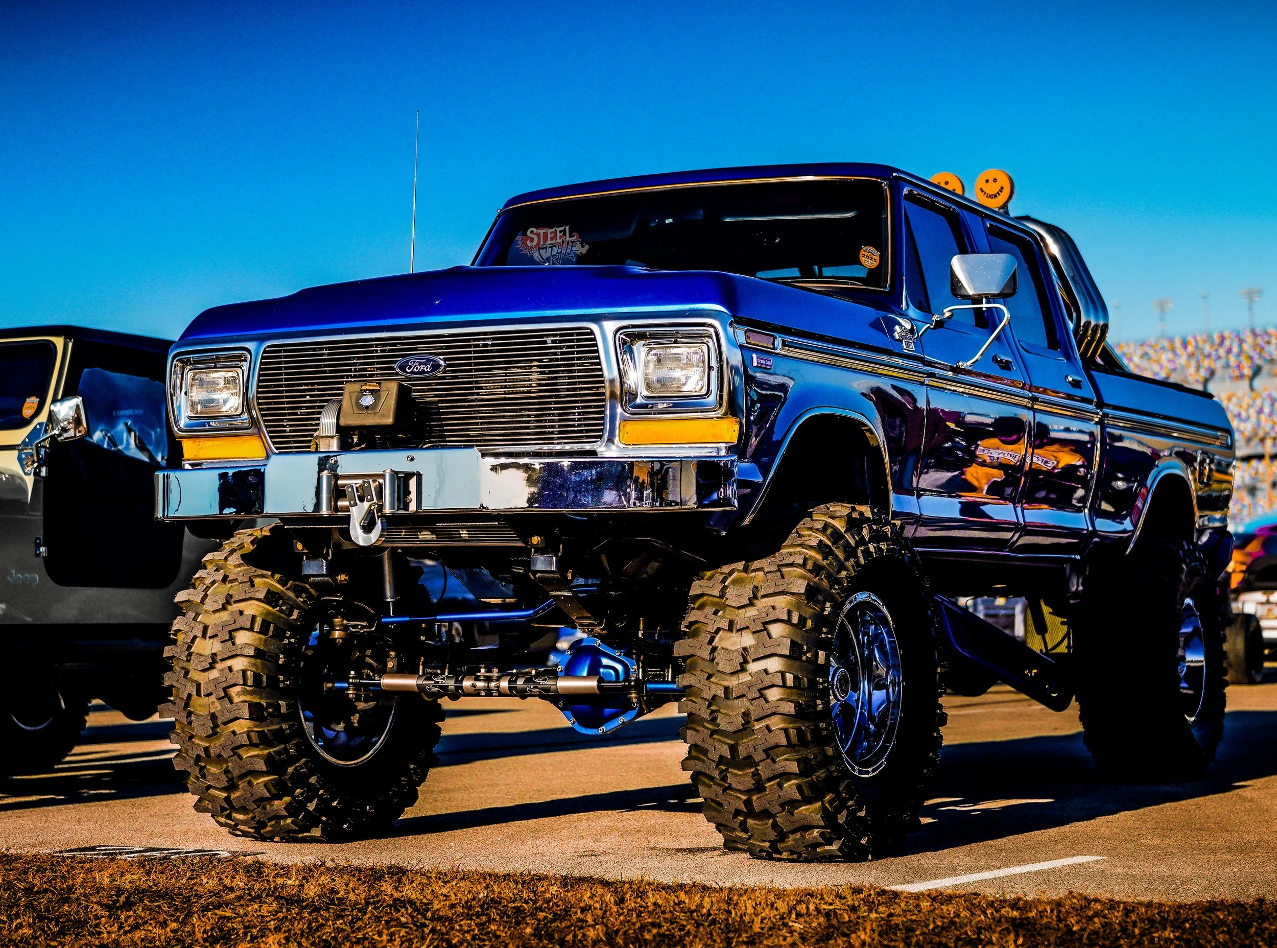 A lifted blue Ford truck with large off-road tires parked on a racetrack during daytime.