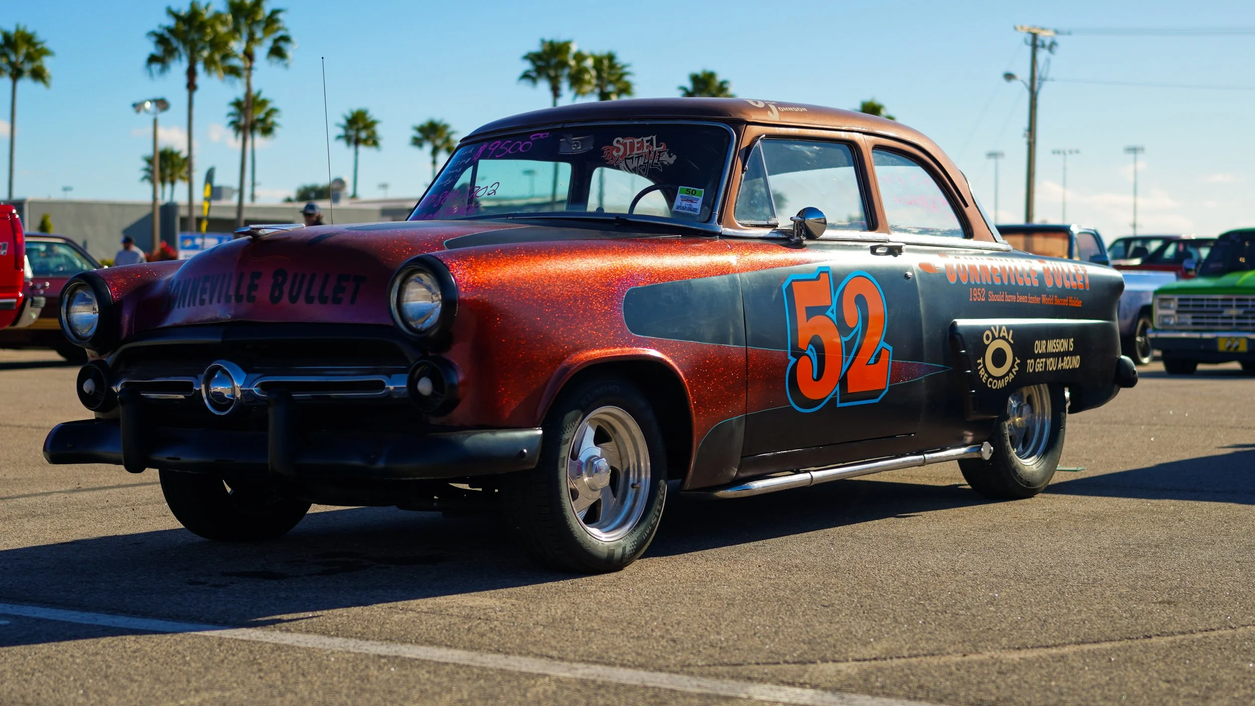 A vintage race car decorated with sponsorship and racing decals, parked in a lot with palm trees in the background.
