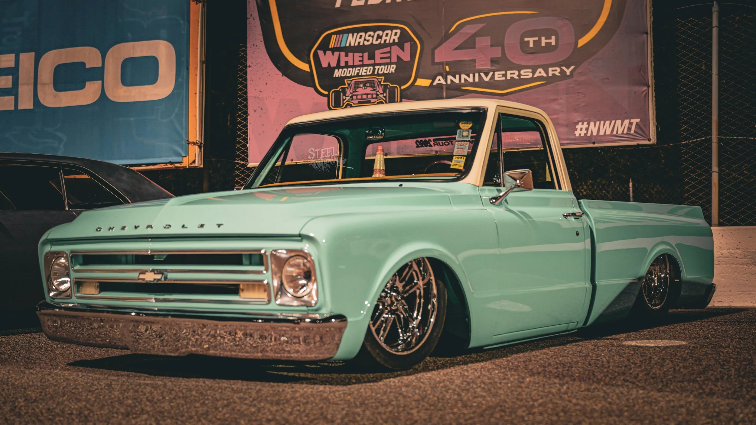 Mint green vintage Chevrolet pickup truck with lowered stance and custom black wheels parked at night with banners in the background celebrating the 40th anniversary of the NASCAR Whelen Modified Tour.