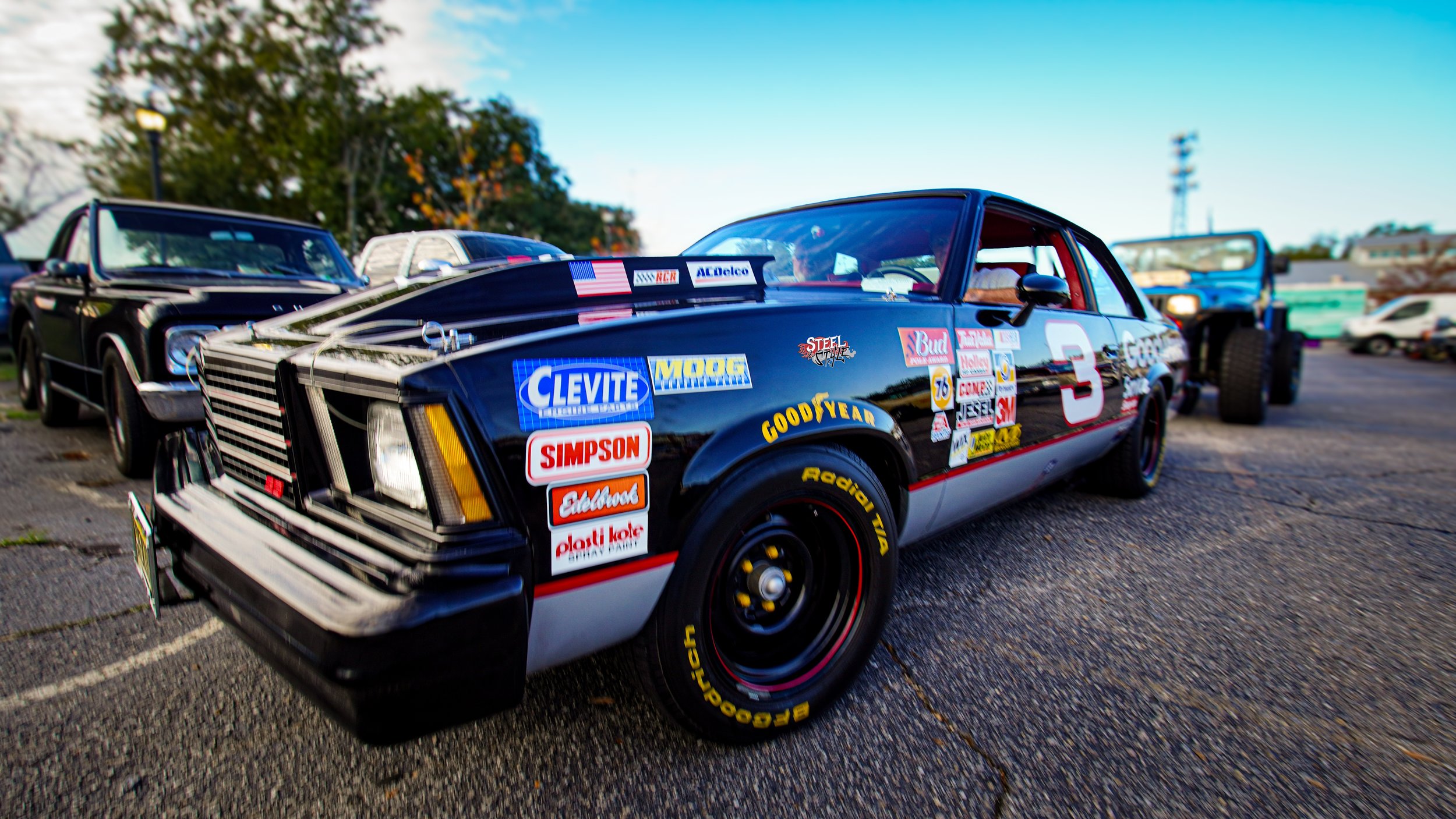 A vintage race car with a black body and various sponsor stickers parked on an outdoor lot, with other classic cars and trucks in the background.