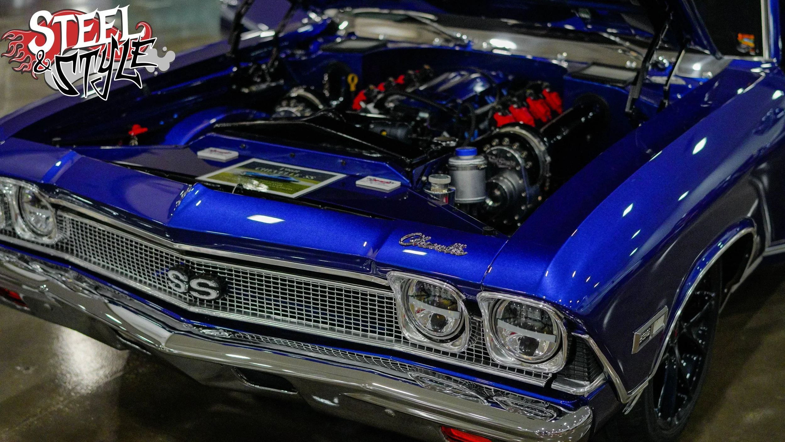 Blue Chevrolet Chevelle SS muscle car with an exposed engine on display, showing the front grille, headlights, and wheels.