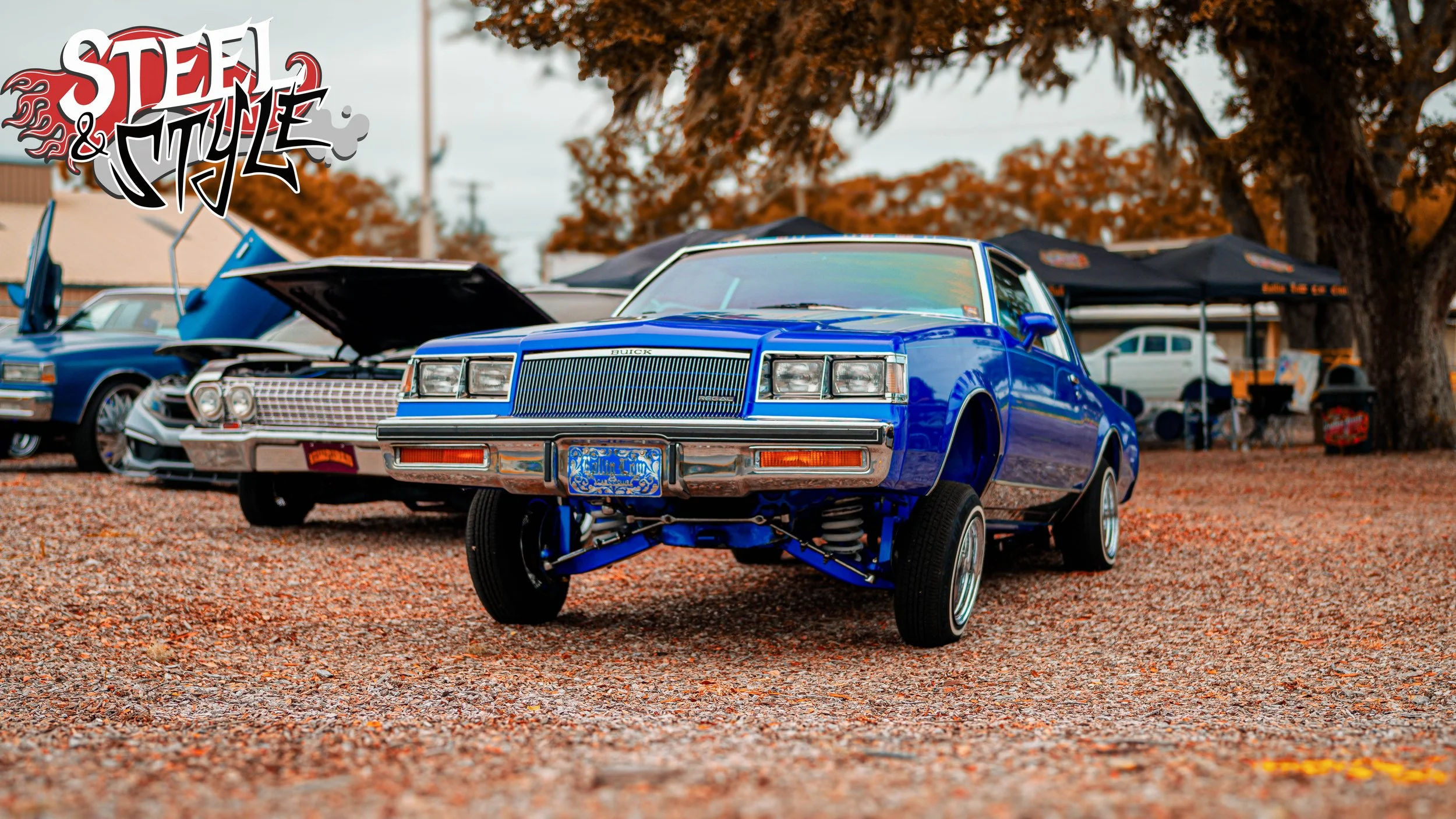 Blue vintage car with its front wheels lifted at a car show, with other classic cars and trees with autumn leaves in the background.