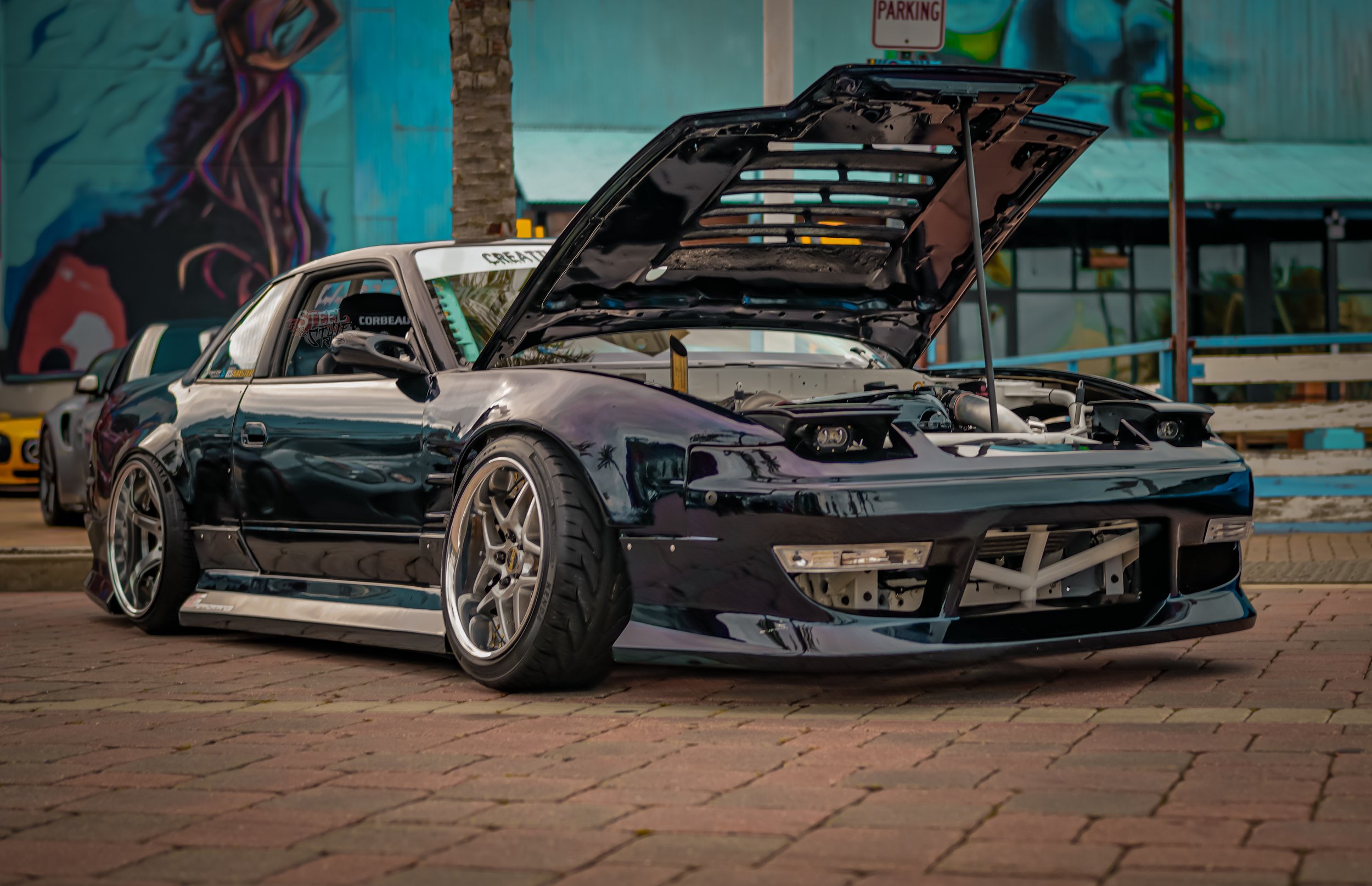 A black sports car with its hood open, parked on a brick surface, showing a racing seat inside and modifications.