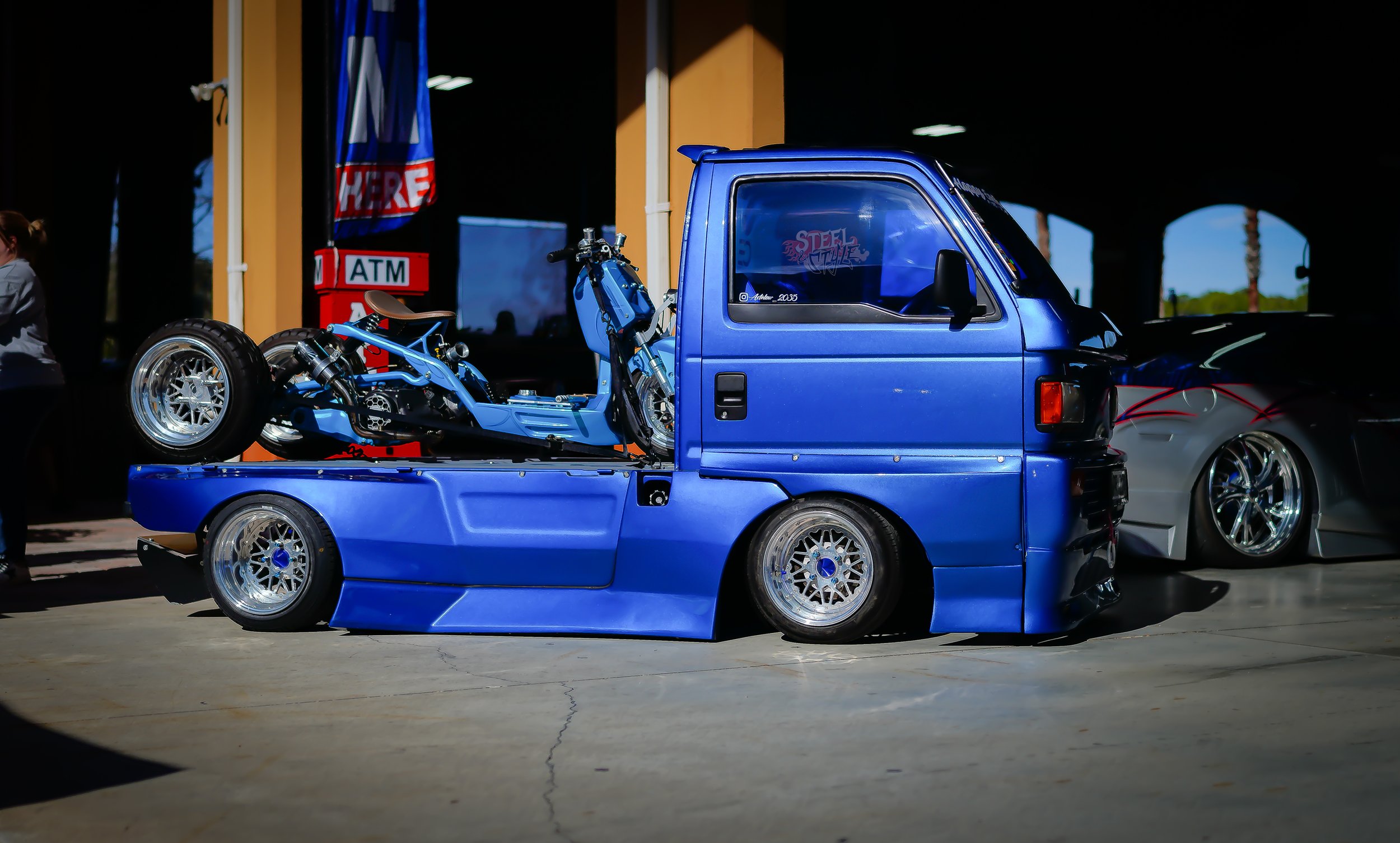 A modified blue mini truck with a motorcycle mounted on its bed, parked indoors at a car show.