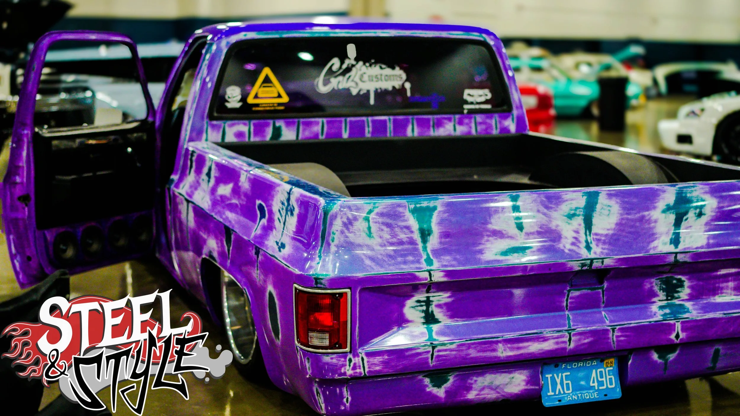 Purple truck with a custom tie-dye paint job, open door, and a Florida license plate that reads 'IX6 496'. The back of the truck is empty, and there are several other cars in the background.
