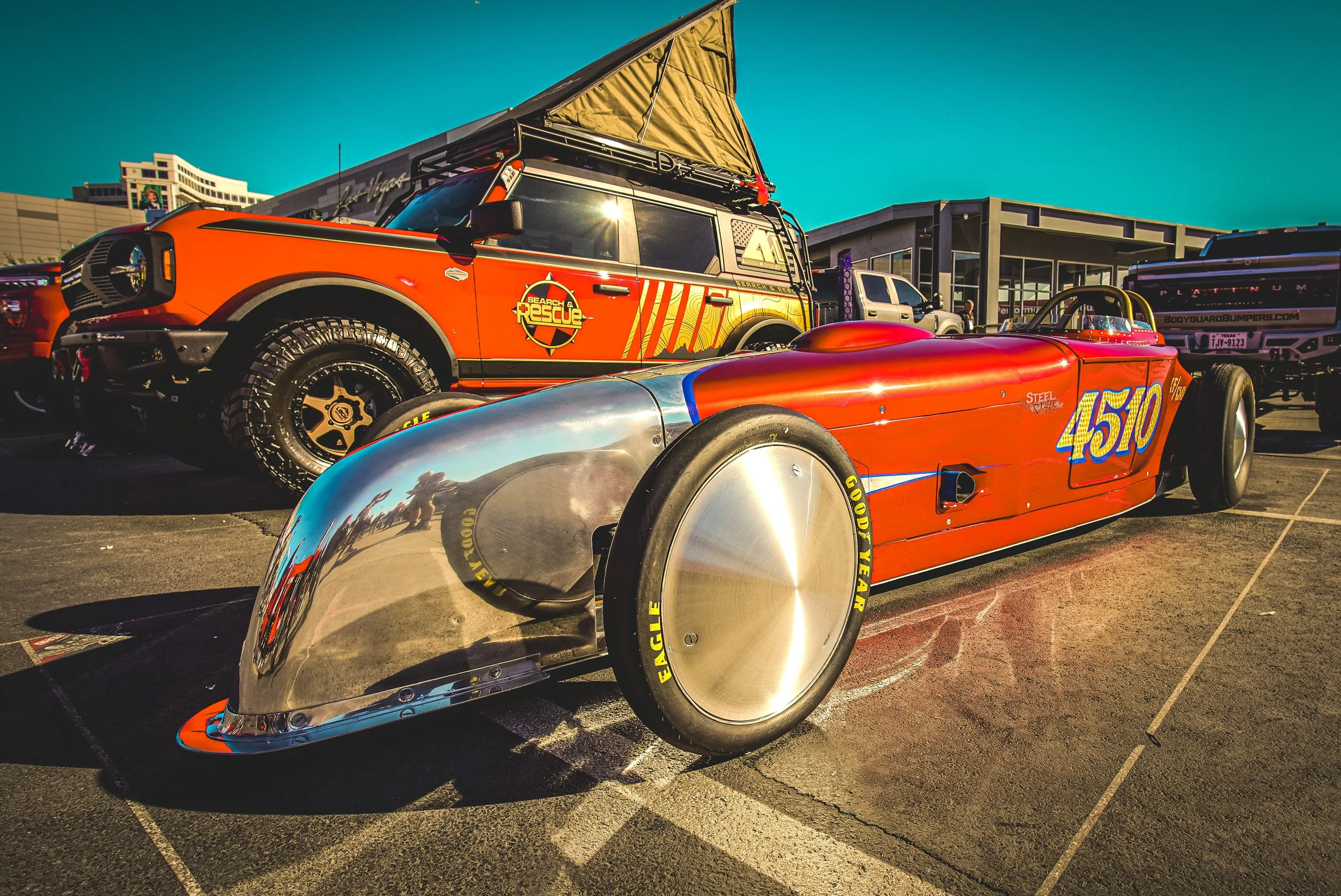 A red vintage racing car with the number 4510, parked outdoors alongside other vehicles, with a building in the background and sunlight reflecting off its chrome parts.