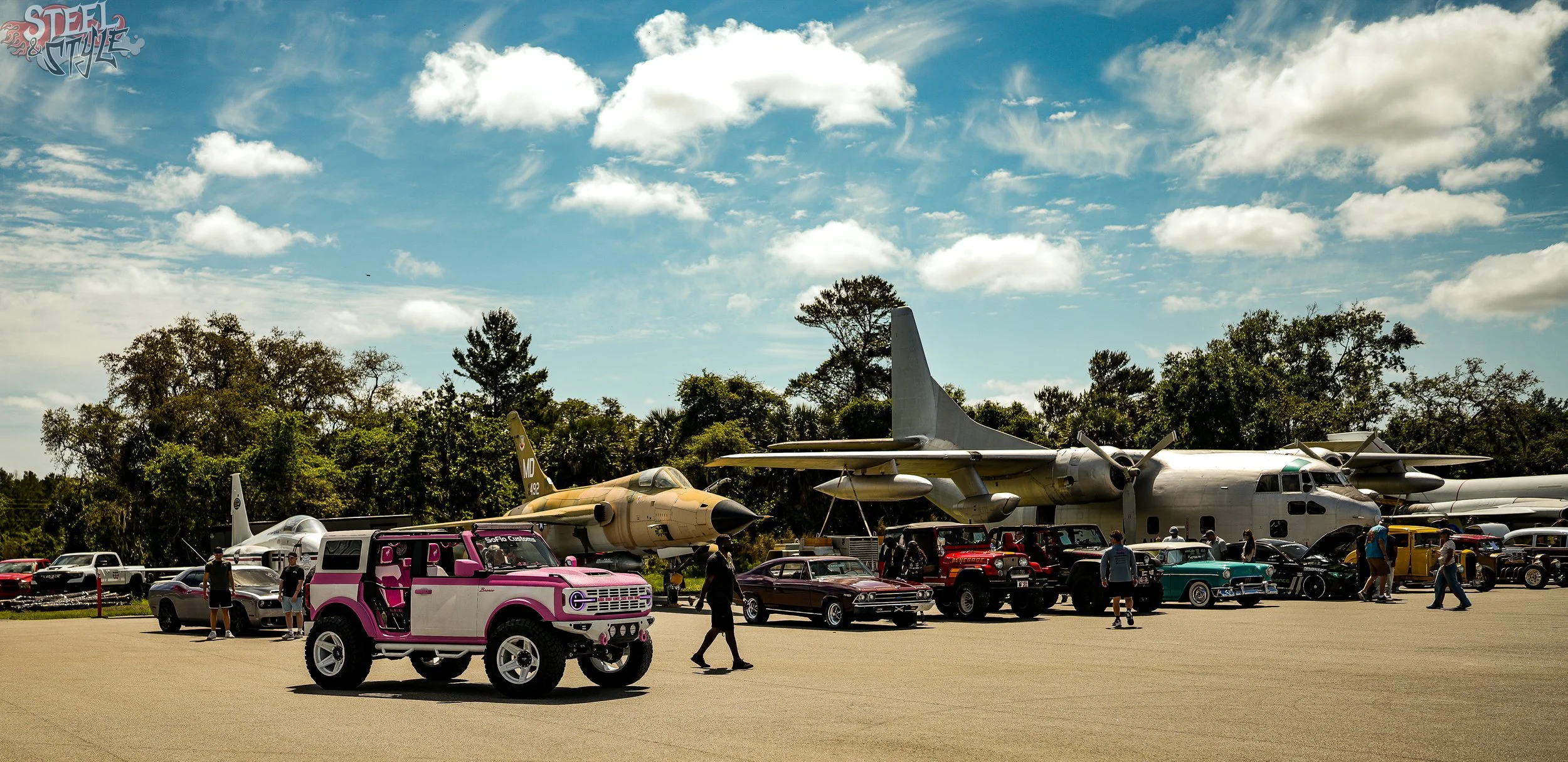 Various classic and military aircraft on display outdoors at an air show, with people walking around and cars parked nearby.