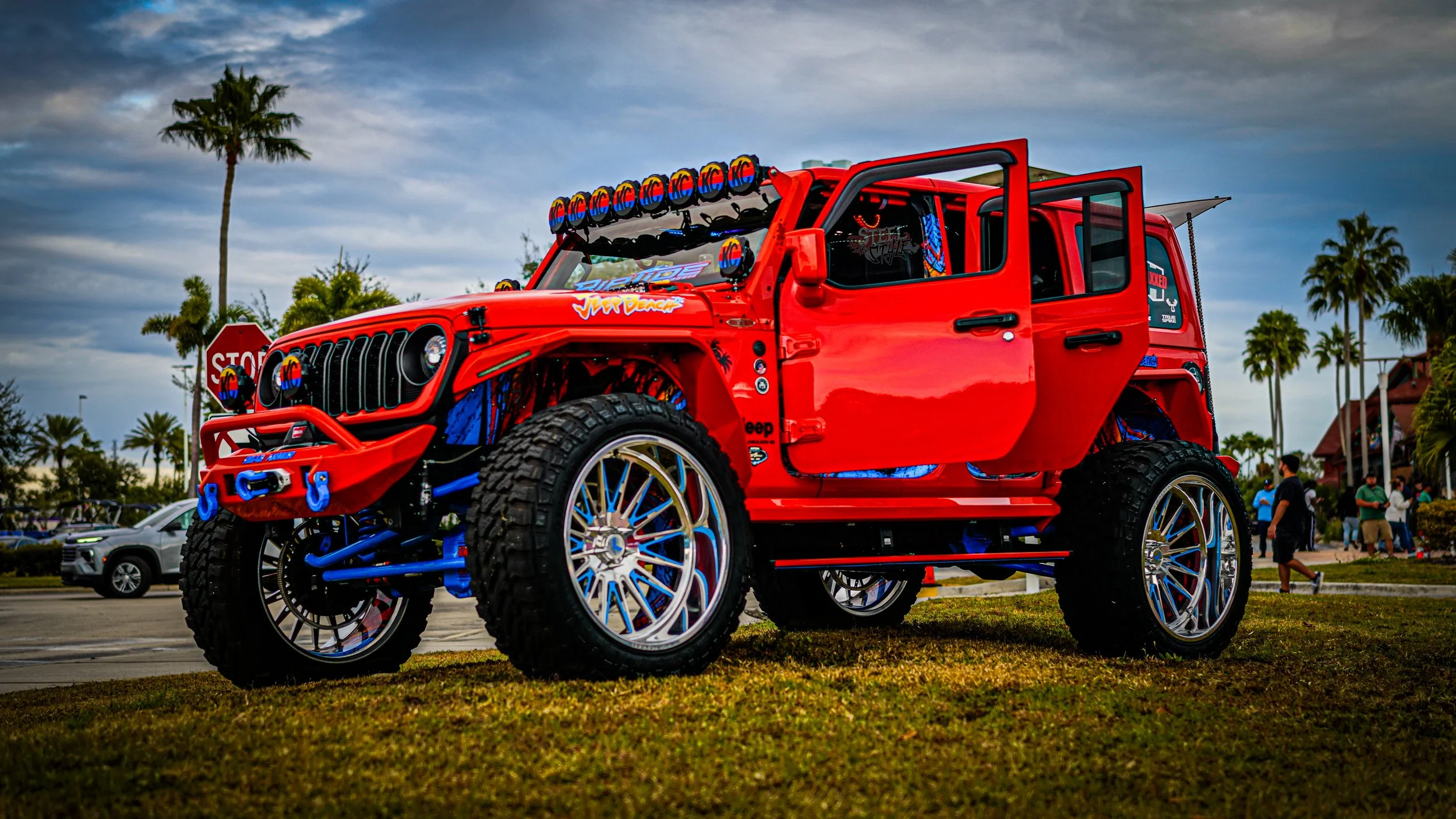 Red customized Jeep with large tires, blue suspension, off-road lights, and decals, parked on grass with palm trees and people in the background.