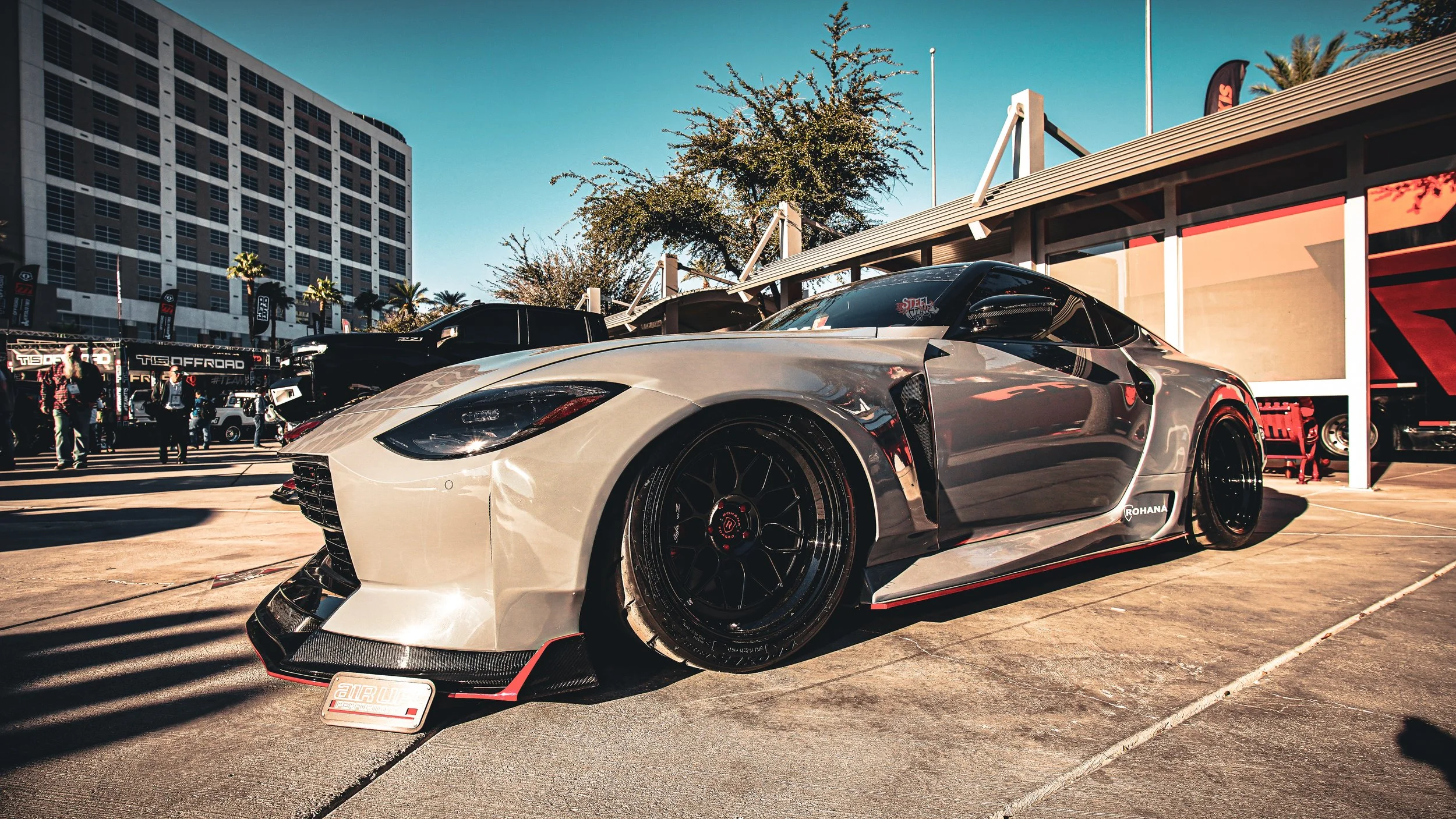 A sleek silver sports car with black wheels parked outdoors at a car event, with people, tents, and a large building in the background.