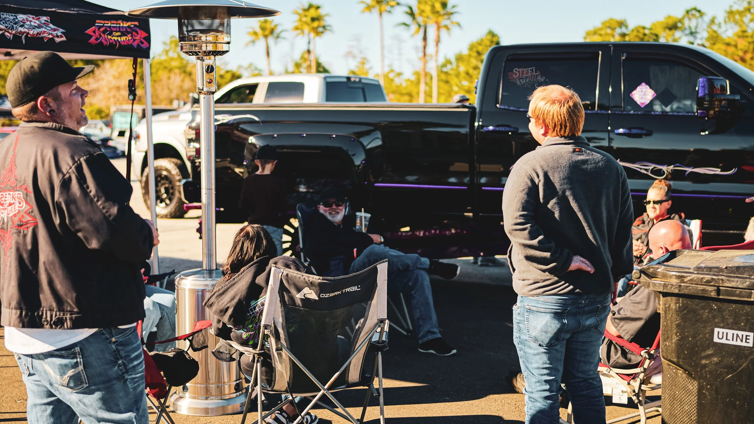 Group of people gathered outdoors around a black pickup truck, some sitting and some standing, with a patio heater and folding chairs, in a parking lot with trees in the background.